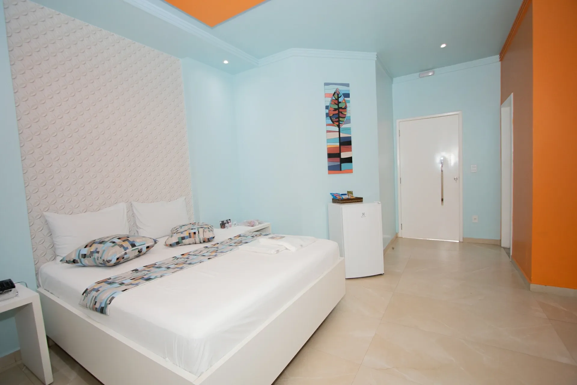 Suíte Hidro Plus do Motel Opium em Boa Vista: Quarto da suíte Hidro: cama de casal box branca com roupa de cama branca e faixa/almofadas em estampa pixelada