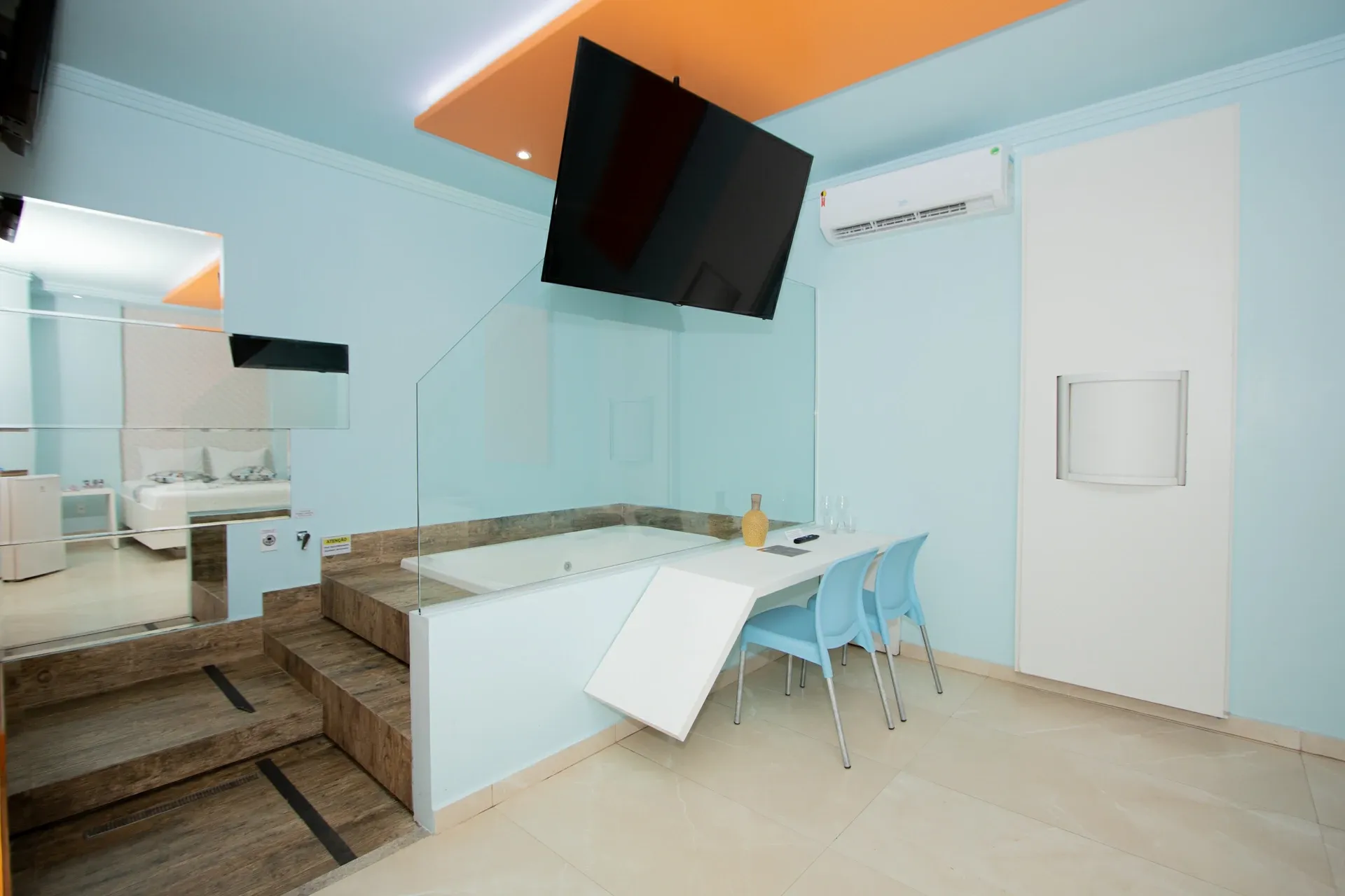 Suíte Hidro Plus do Motel Opium em Boa Vista: Mesmo enquadramento de HIDRO-6: banheira no deque amadeirado atrás do vidro, TV suspensa, bancada com cadeiras