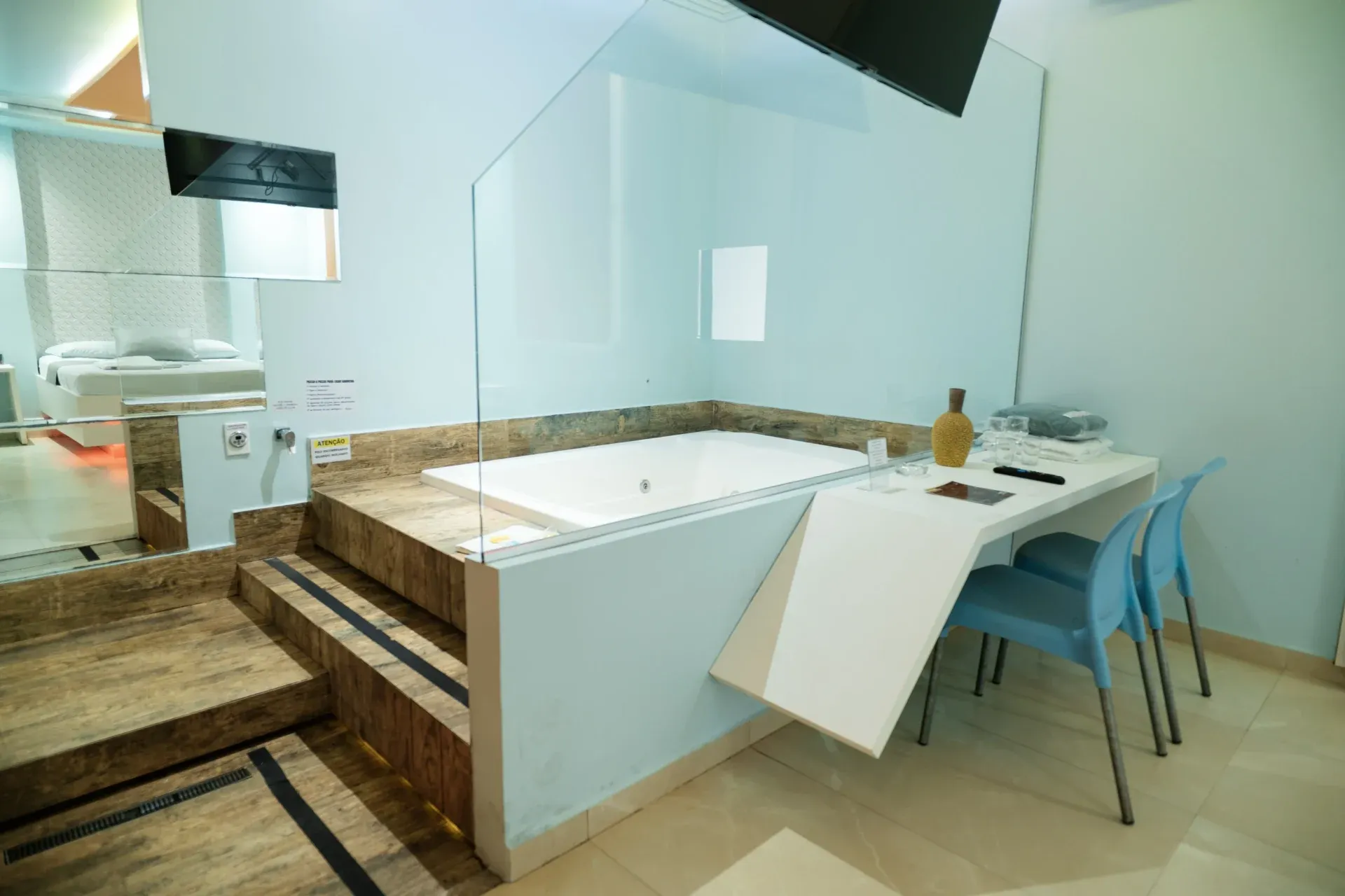 Suíte Hidro Plus do Motel Opium em Boa Vista: Suíte clara em azul-pastel com banheira de hidromassagem branca embutida em plataforma elevada revestida de po