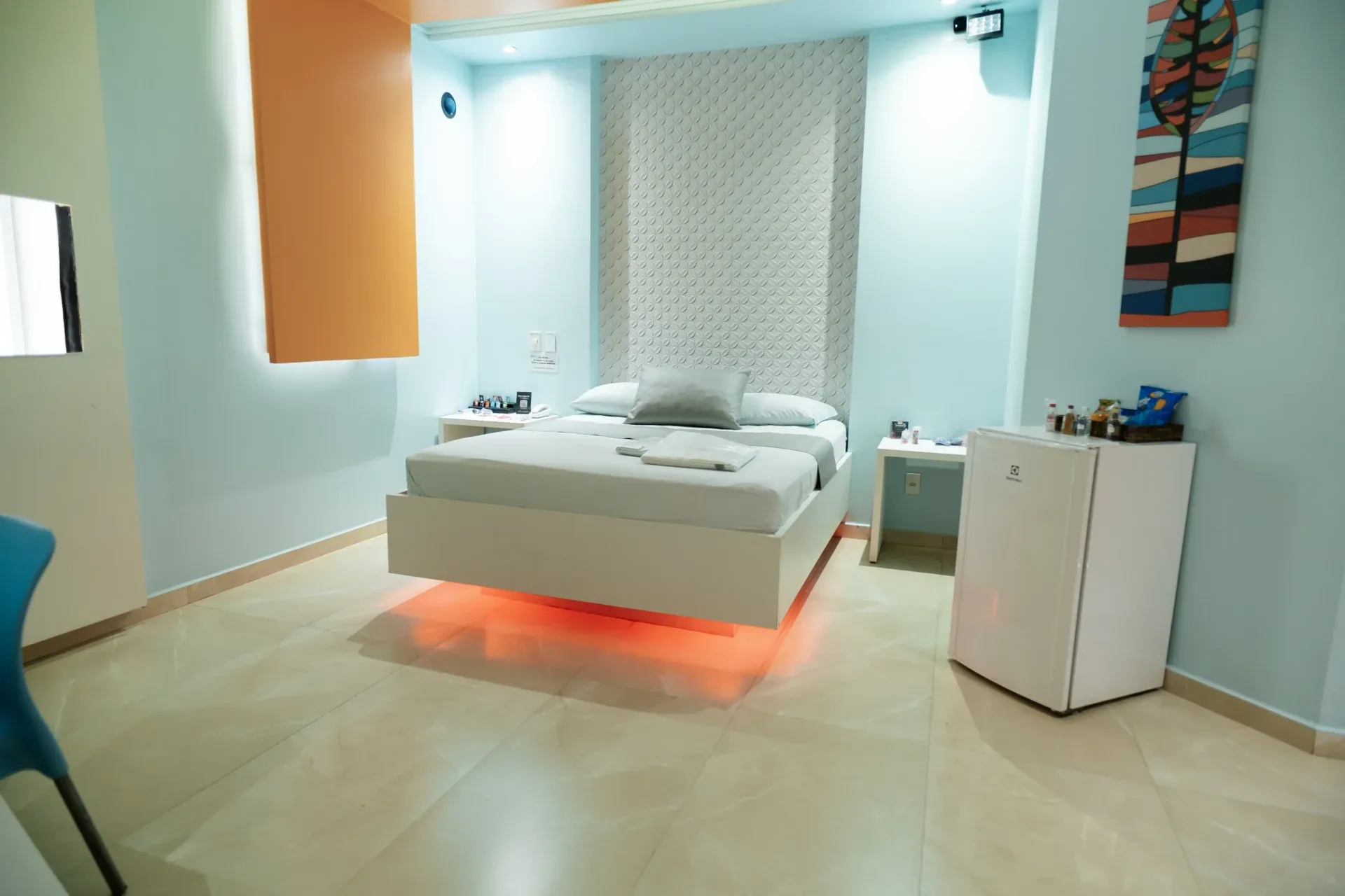 Suíte Hidro Plus do Motel Opium em Boa Vista: Quarto claro em azul-pastel com cama 'flutuante' branca sobre LED laranja (cromoterapia), painel 3D branco tex