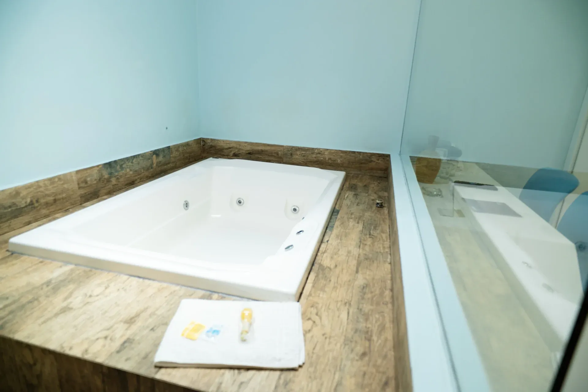 Suíte Hidro Plus do Motel Opium em Boa Vista: Close em ângulo alto da banheira de hidromassagem branca retangular embutida em deck de porcelanato amadeirado