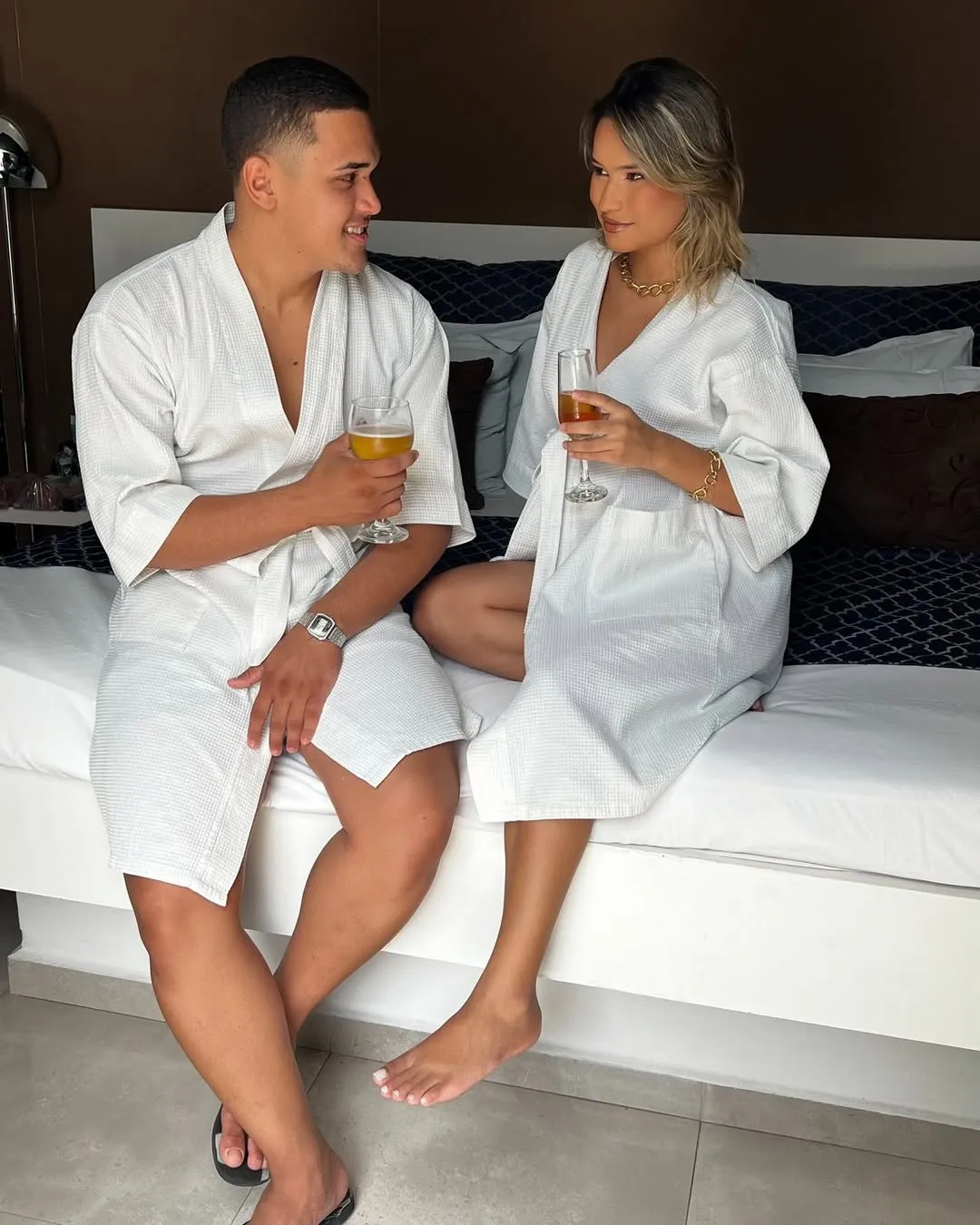 Motel Opium: Conteúdo lifestyle de rede social: casal sentado na beirada de cama box branca vestindo roupões brancos atoalh
