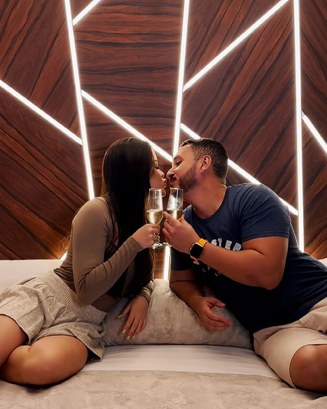 Motel Opium: 5) de um casal sentado na cama brindando com taças de espumante prestes a se beijar, diante de cabeceira-paine