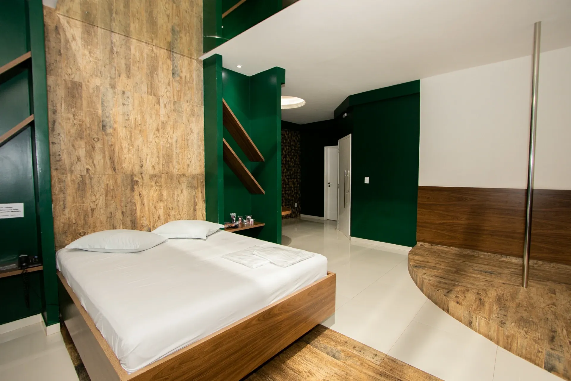 Suíte Luxo do Motel Opium em Boa Vista: Suíte com paredes verde-esmeralda e painel de madeira rústica atrás da cama box de madeira com roupa branca