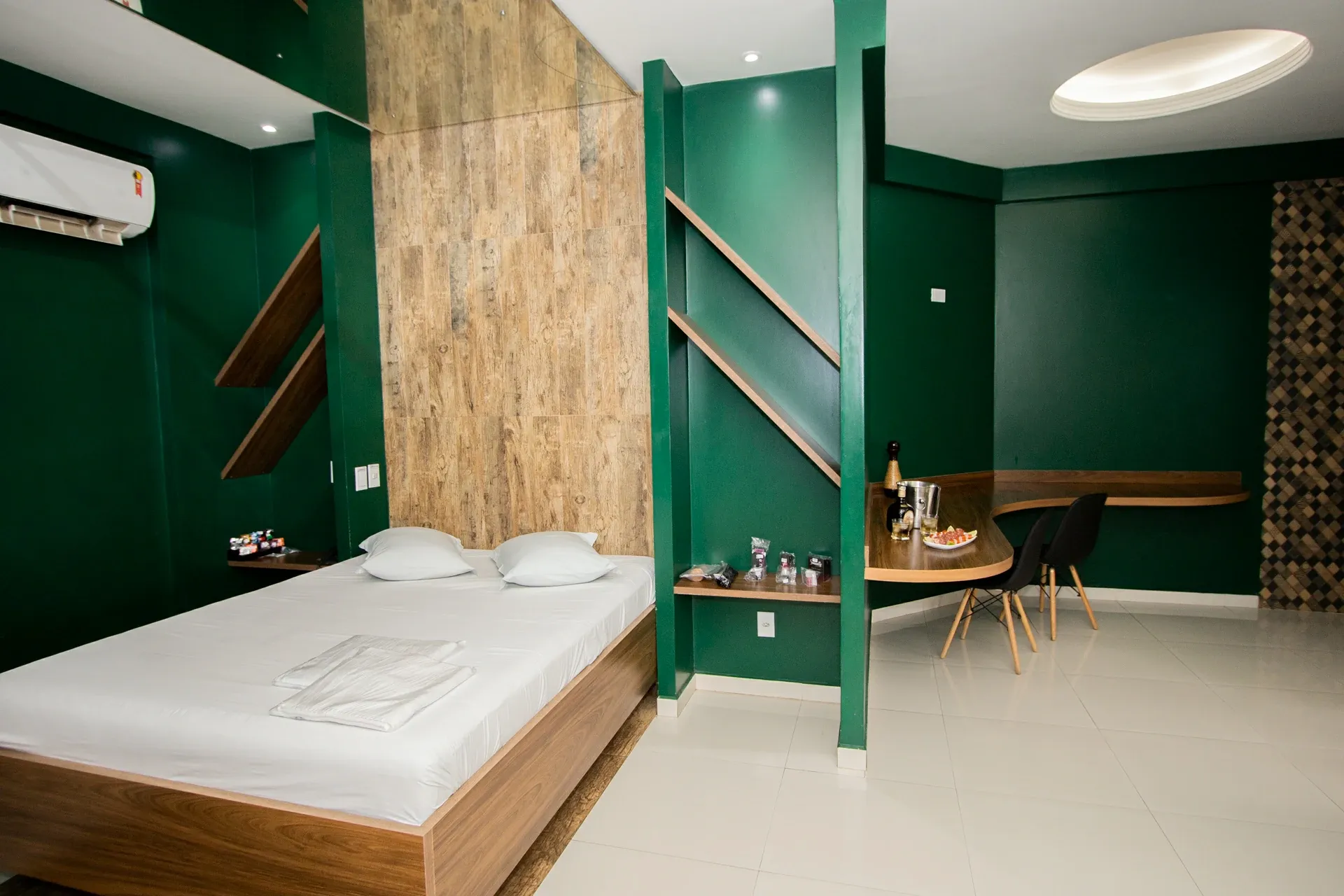 Suíte Luxo do Motel Opium em Boa Vista: Mesma suíte verde vista do outro ângulo: cama à esquerda, bancada curva de madeira com balde de gelo, bebida e