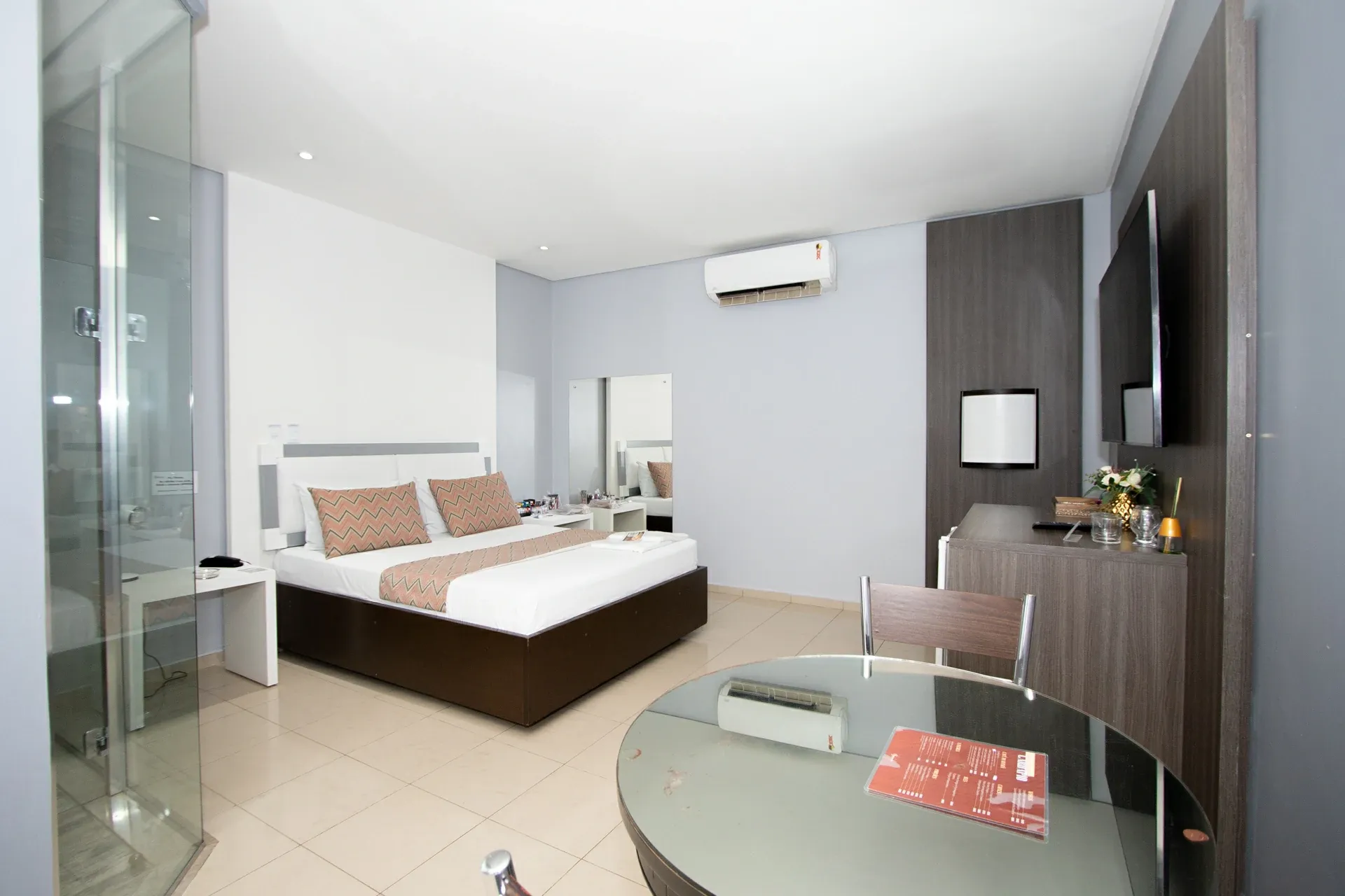 Suíte Luxo do Motel Opium em Boa Vista: Suíte clara em cinza e branco com cama de base em madeira escura, almofadas chevron salmão, painel de madeira 