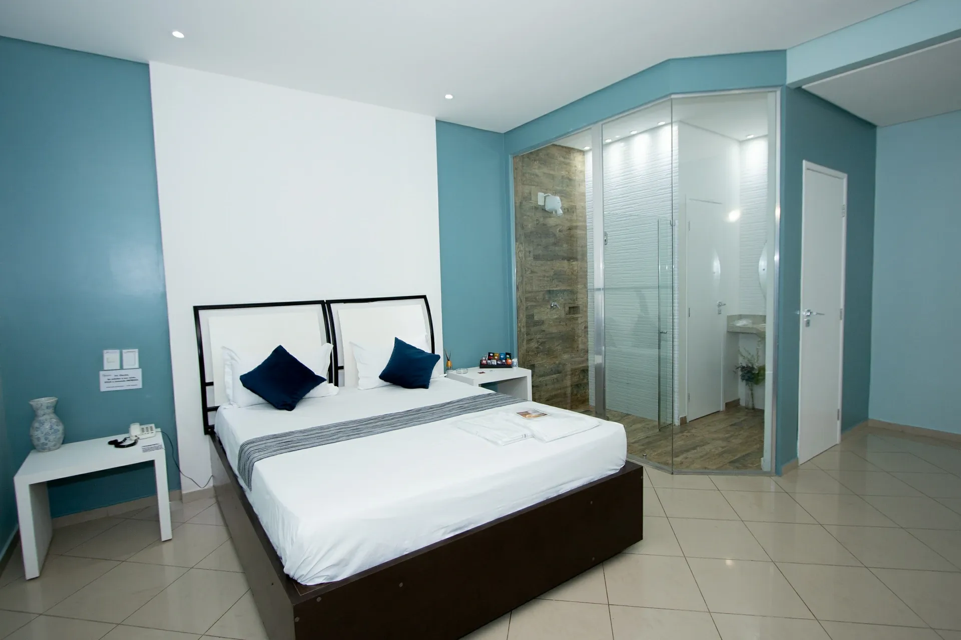 Suíte Luxo do Motel Opium em Boa Vista: Suíte com paredes azul-petróleo e parede branca de destaque, cama com cabeceiras duplas brancas e almofadas az