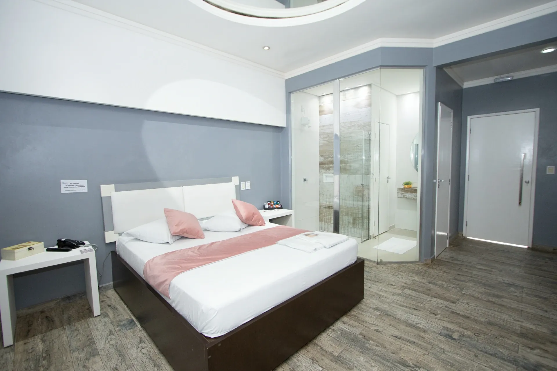 Suíte Luxo do Motel Opium em Boa Vista: Suíte cinza-azulada com cabeceira estofada branca, almofadas e manta rosé, piso vinílico/porcelanato amadeirad