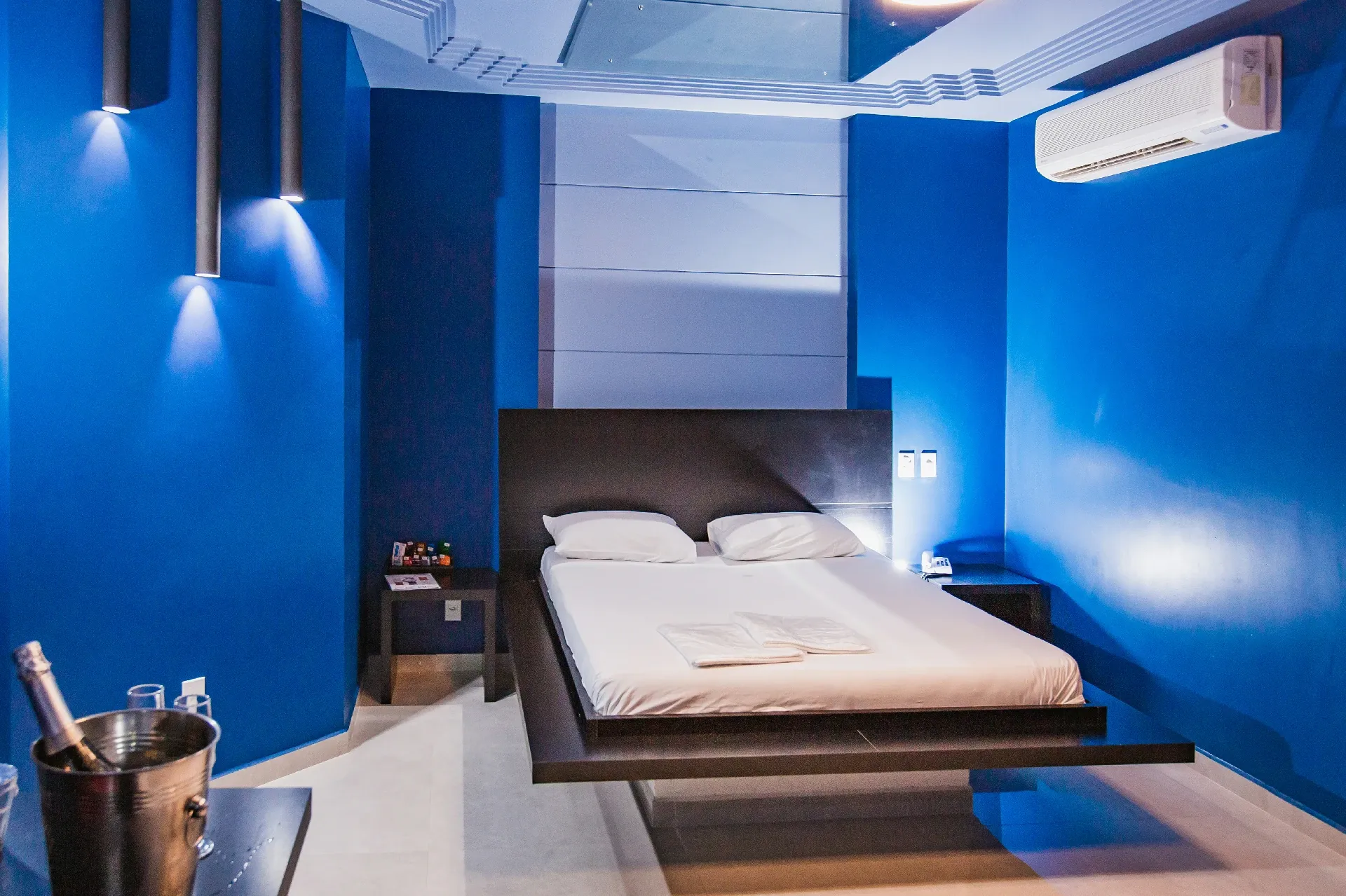 Suíte Luxo do Motel Opium em Boa Vista: Suíte azul-cobalto com iluminação cênica: cama plataforma flutuante de madeira escura sob espelho de teto, spo