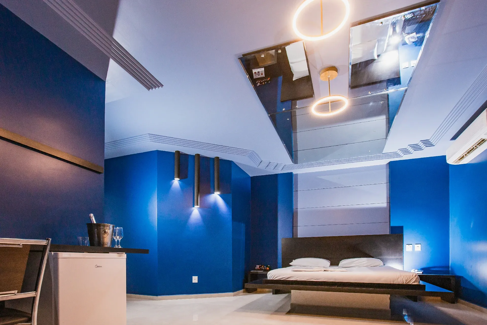 Suíte Luxo do Motel Opium em Boa Vista: Mesma suíte azul-cobalto em ângulo amplo mostrando o pé-direito alto: grande espelho de teto com duas luminári
