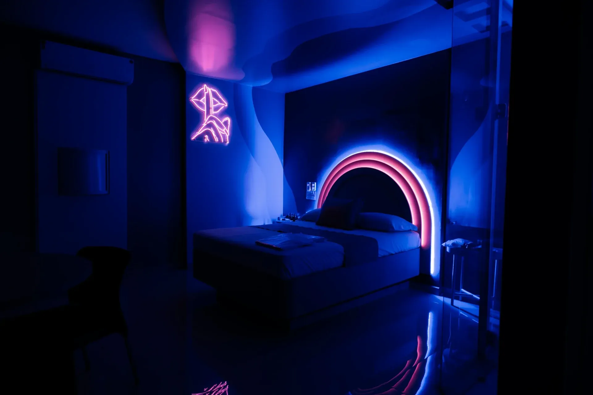 Suíte Luxo do Motel Opium em Boa Vista: Cama casal diante de cabeceira arqueada contornada por LED rosa/branco, neon rosa na parede em forma de silhue