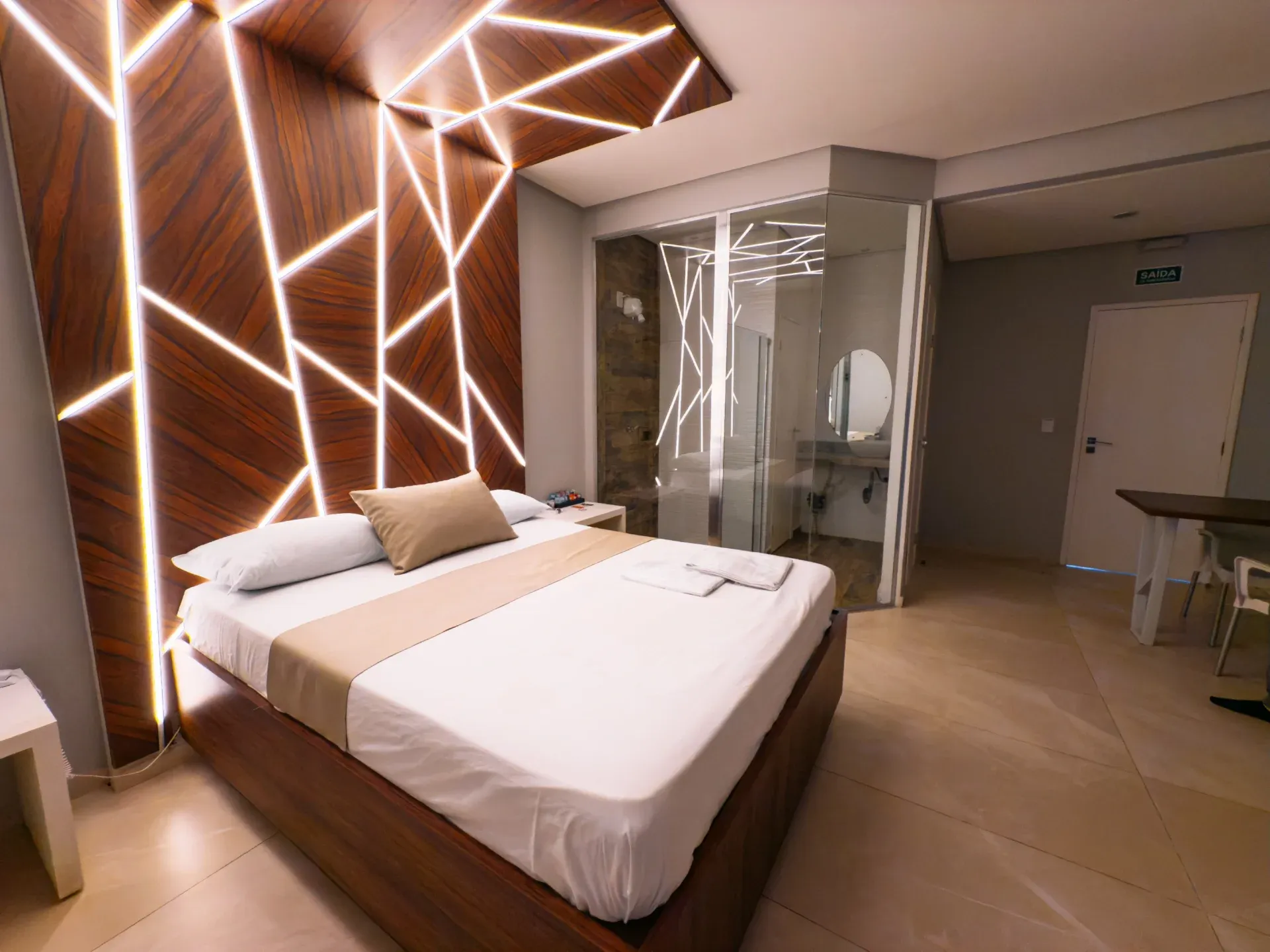 Suíte Luxo do Motel Opium em Boa Vista: Suíte com cama de casal em base de madeira e uma parede-cabeceira escultural de painéis amadeirados cortados p