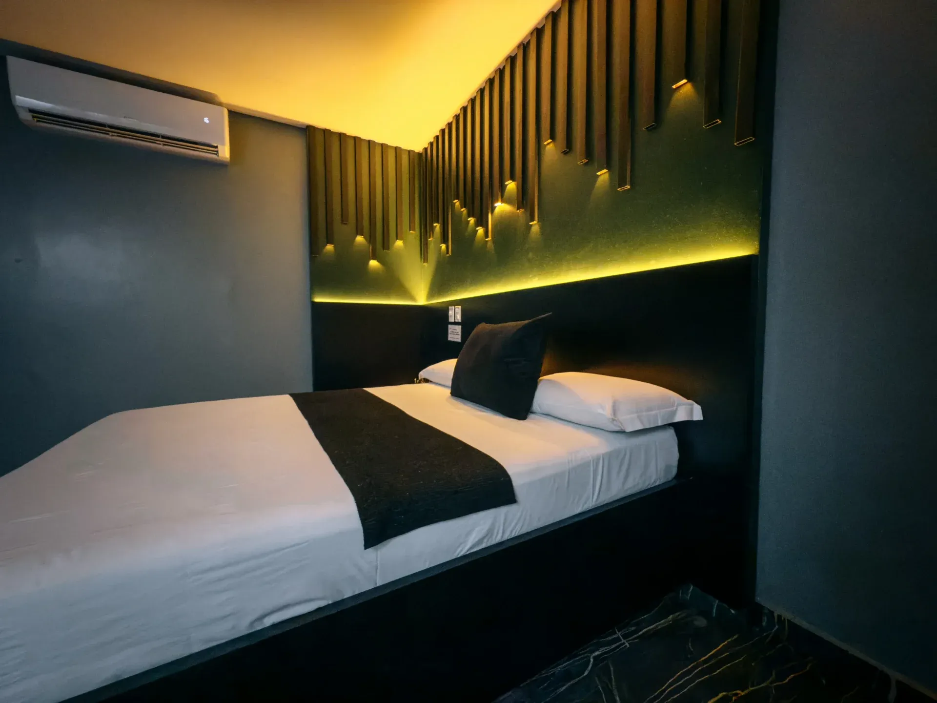 Suíte Luxo do Motel Opium em Boa Vista: Painel de ripas verticais escuras com mini-arandelas embutidas e fita de LED dourada indireta sobre cabeceira 