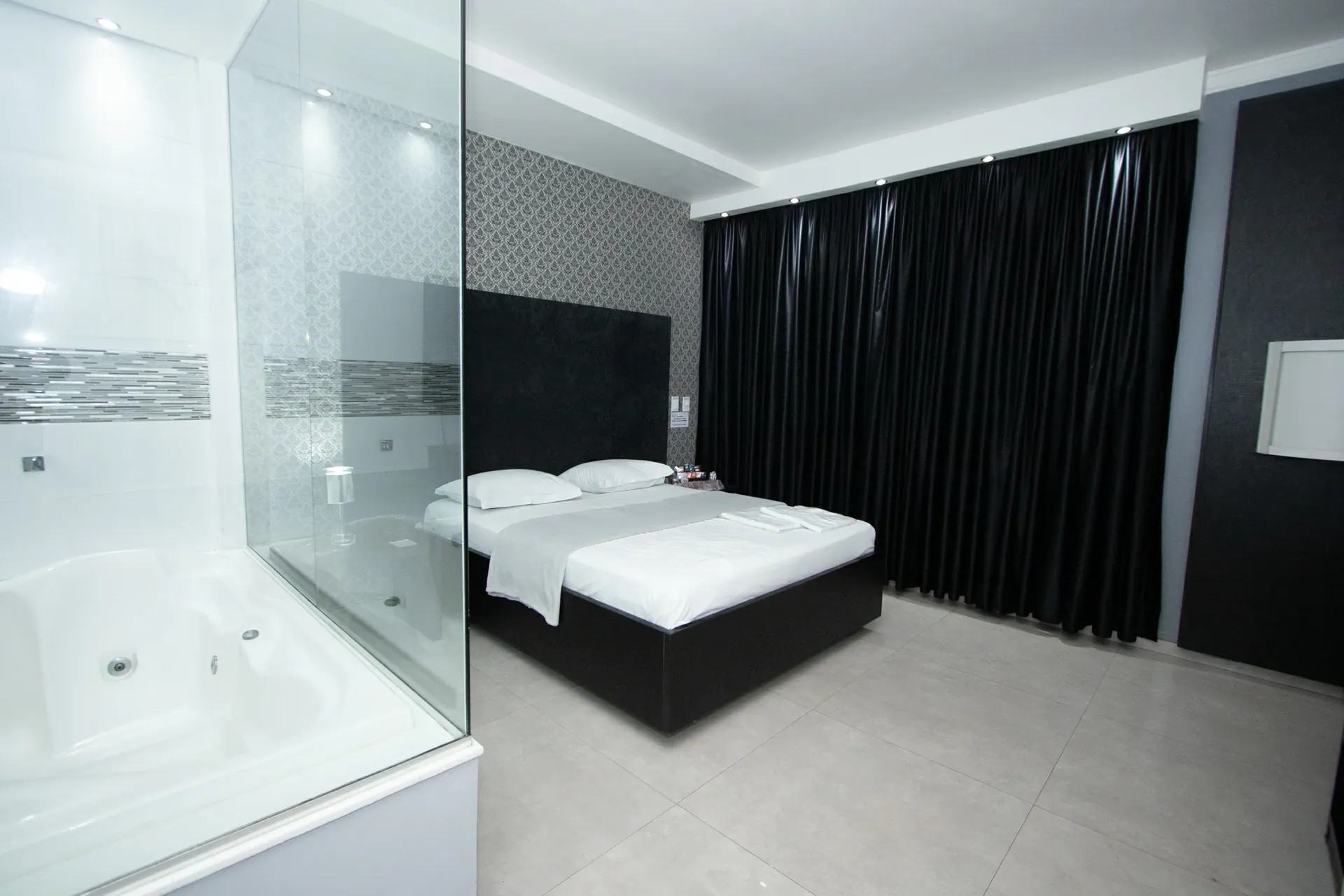 Suíte Luxo Hidro do Motel Opium em Boa Vista: Suíte em preto e branco vista da lateral: banheira de hidromassagem branca atrás de divisória de vidro no prim