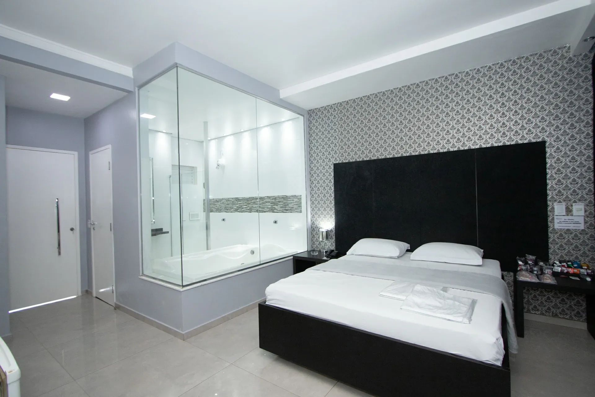 Suíte Luxo Hidro do Motel Opium em Boa Vista: Mesma suíte preto/branco em plano geral: cubo de vidro central com banheira de hidromassagem e box iluminado, 