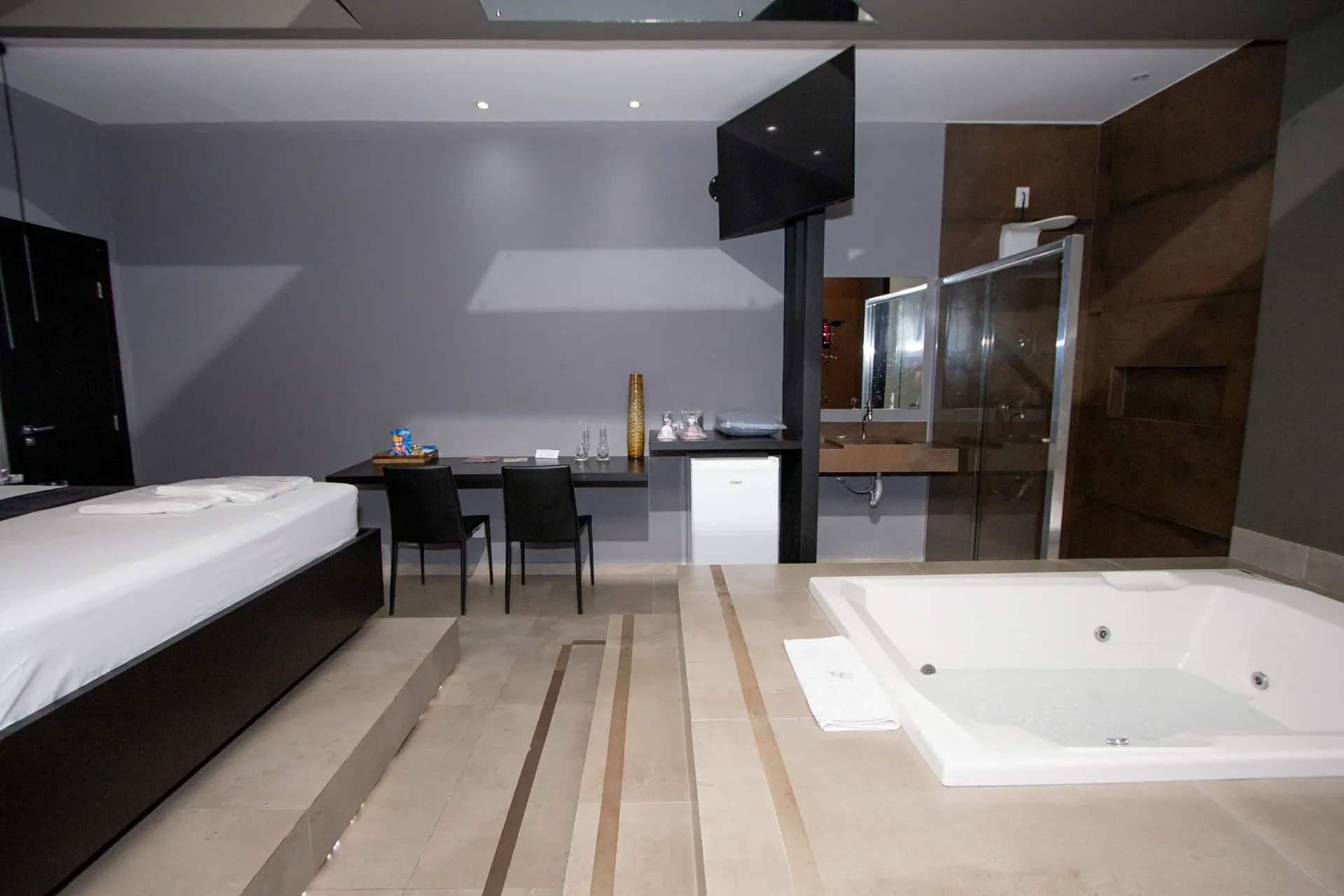 Suíte Luxo Hidro do Motel Opium em Boa Vista: Mesma suíte bege/espelhada no ângulo oposto: cama à esquerda, bancada preta com cadeiras, frigobar e vaso deco