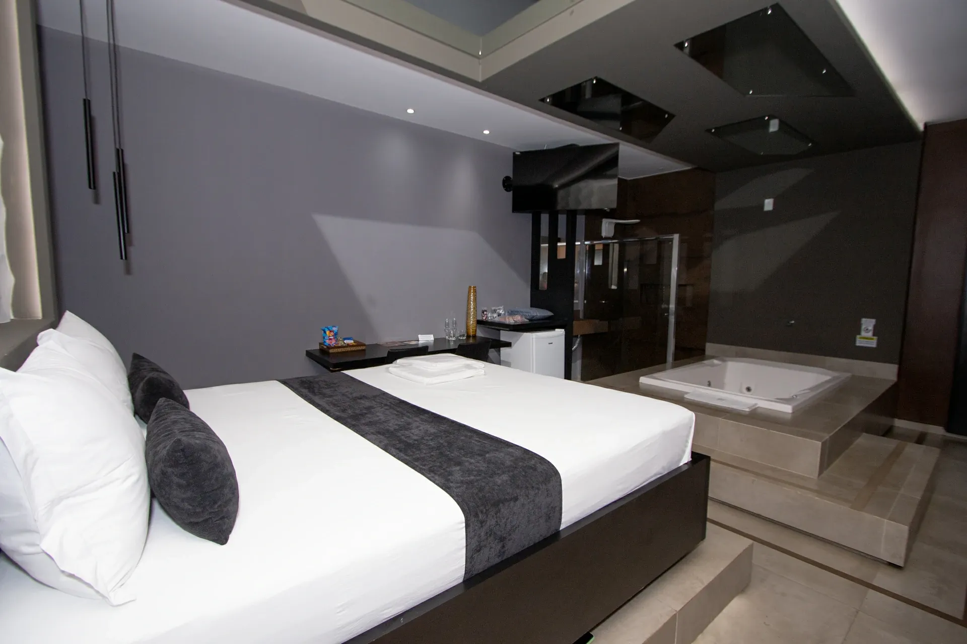 Suíte Luxo Hidro do Motel Opium em Boa Vista: Roupa de cama branca com peseira e almofadas de veludo grafite, pendentes tubulares pretos, teto espelhado com