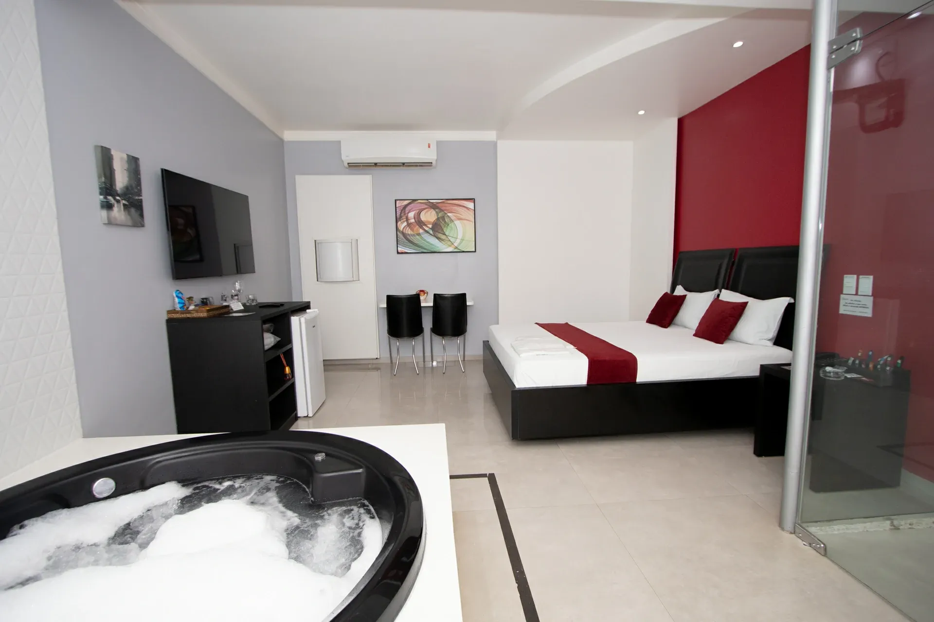 Suíte Luxo Hidro do Motel Opium em Boa Vista: Suíte com parede de destaque vermelha: cama de casal com cabeceira dupla de couro preto, faixa e almofadas de 