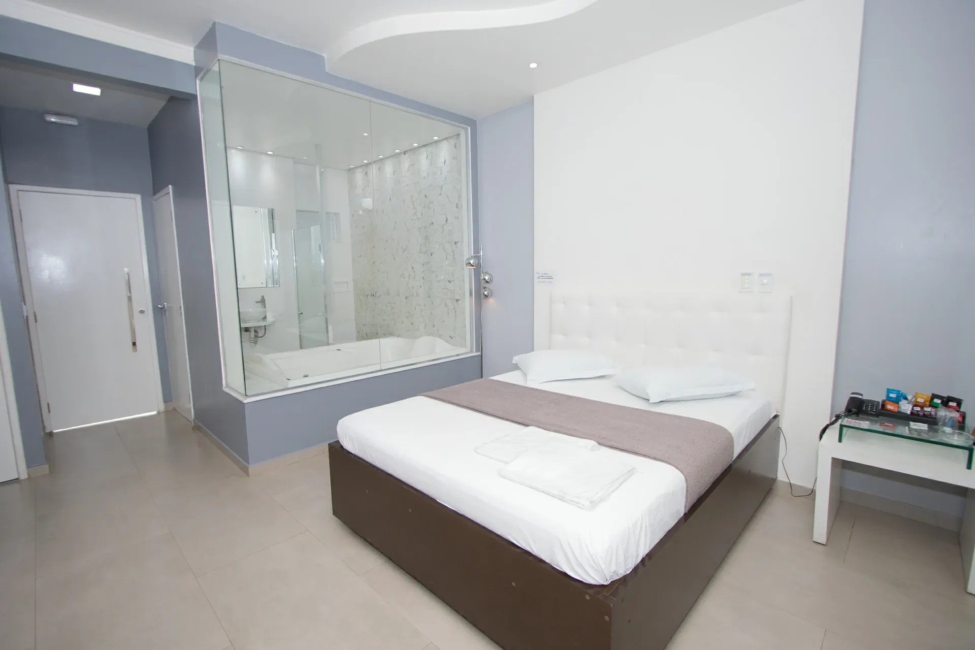 Suíte Luxo Hidro do Motel Opium em Boa Vista: Suíte clara em azul-acinzentado e branco: cama box marrom-escuro com cabeceira capitonê branca e manta taupe, 