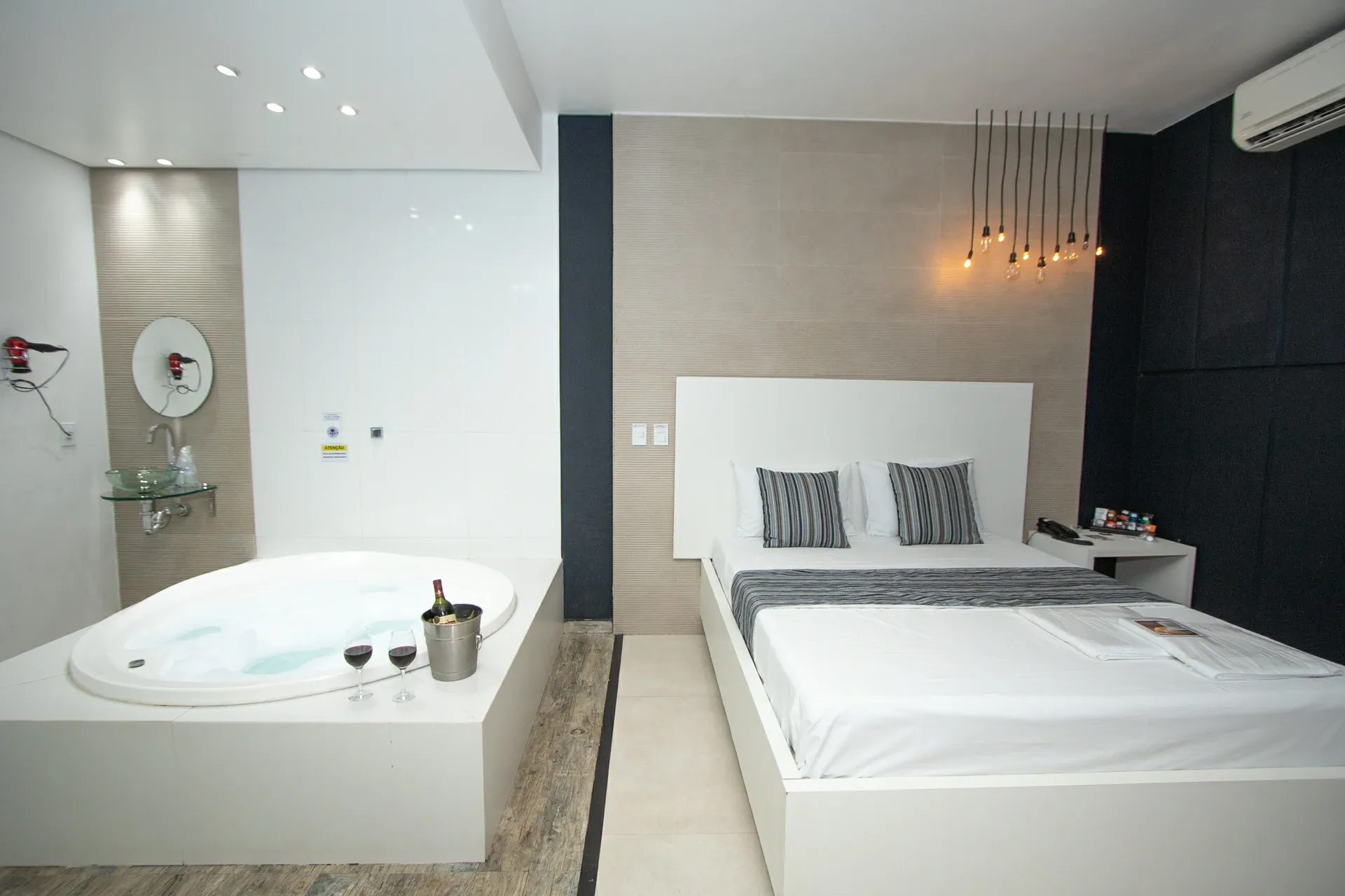 Suíte Luxo Hidro do Motel Opium em Boa Vista: Cama box branca de casal com cabeceira branca e almofadas listradas, e ao lado, sobre plataforma revestida de 