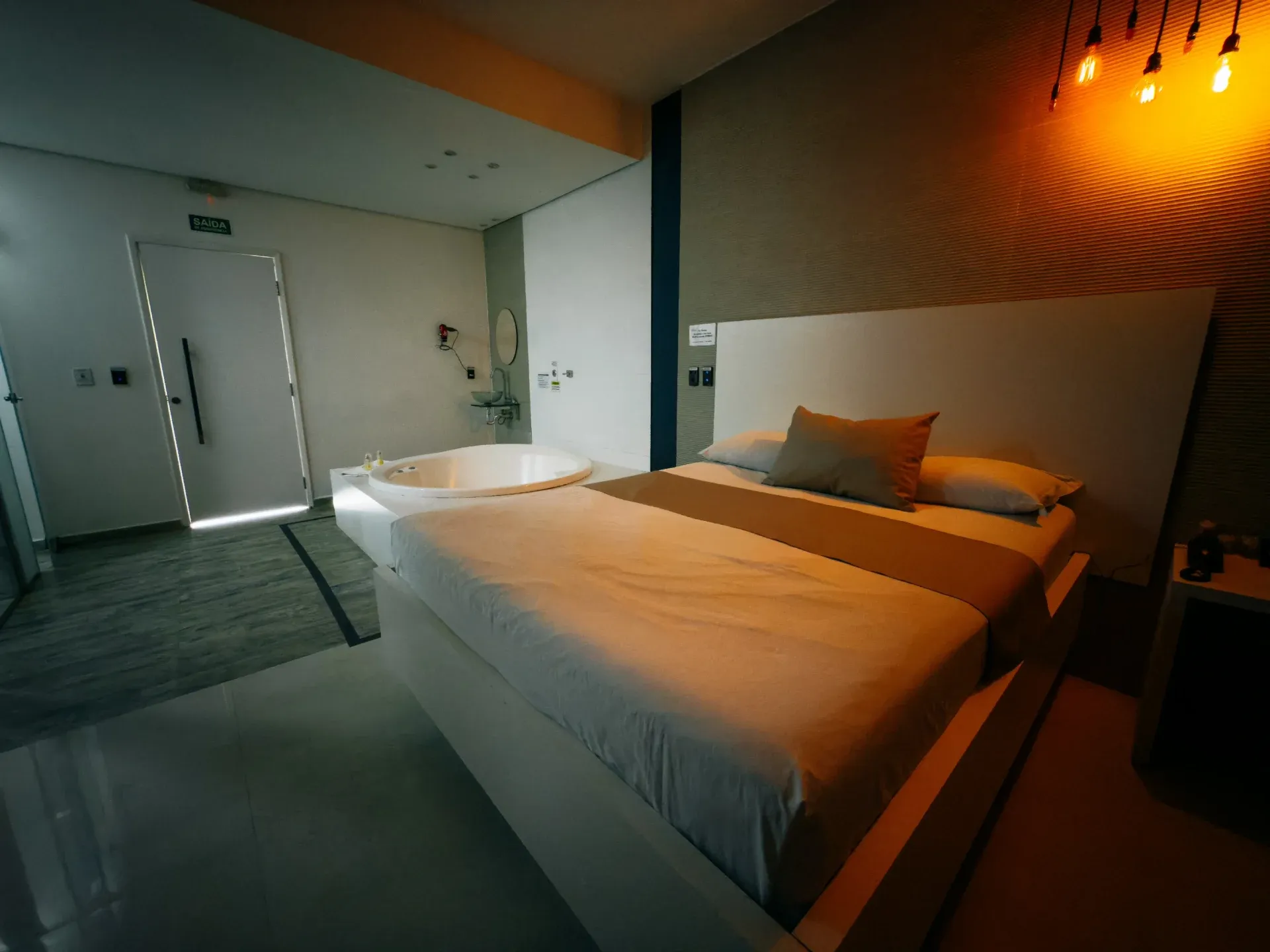 Suíte Luxo Hidro do Motel Opium em Boa Vista: Suíte clara com cama de casal em base branca e cabeceira ampla branca, roupa de cama bege/marrom, hidromassage