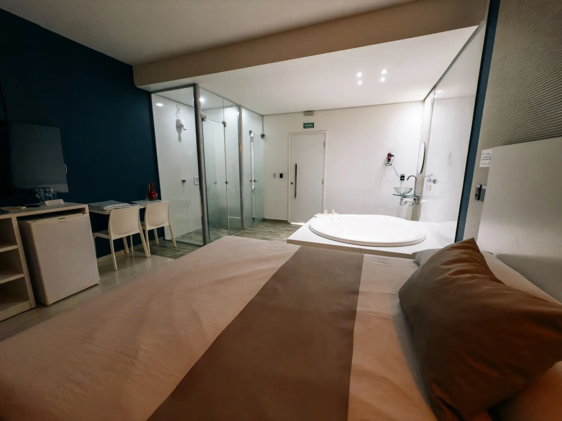 Suíte Luxo Hidro do Motel Opium em Boa Vista: Mesma linha da suíte azul-marinho vista de cima da cama: primeiro plano dominado pela roupa de cama bege/marro