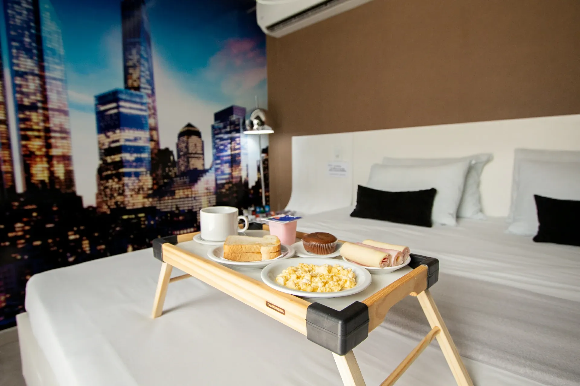 Suíte Opium do Motel Opium em Boa Vista: Bandeja de café da manhã sobre cama de casal branca em suíte com papel de parede fotográfico de skyline noturn