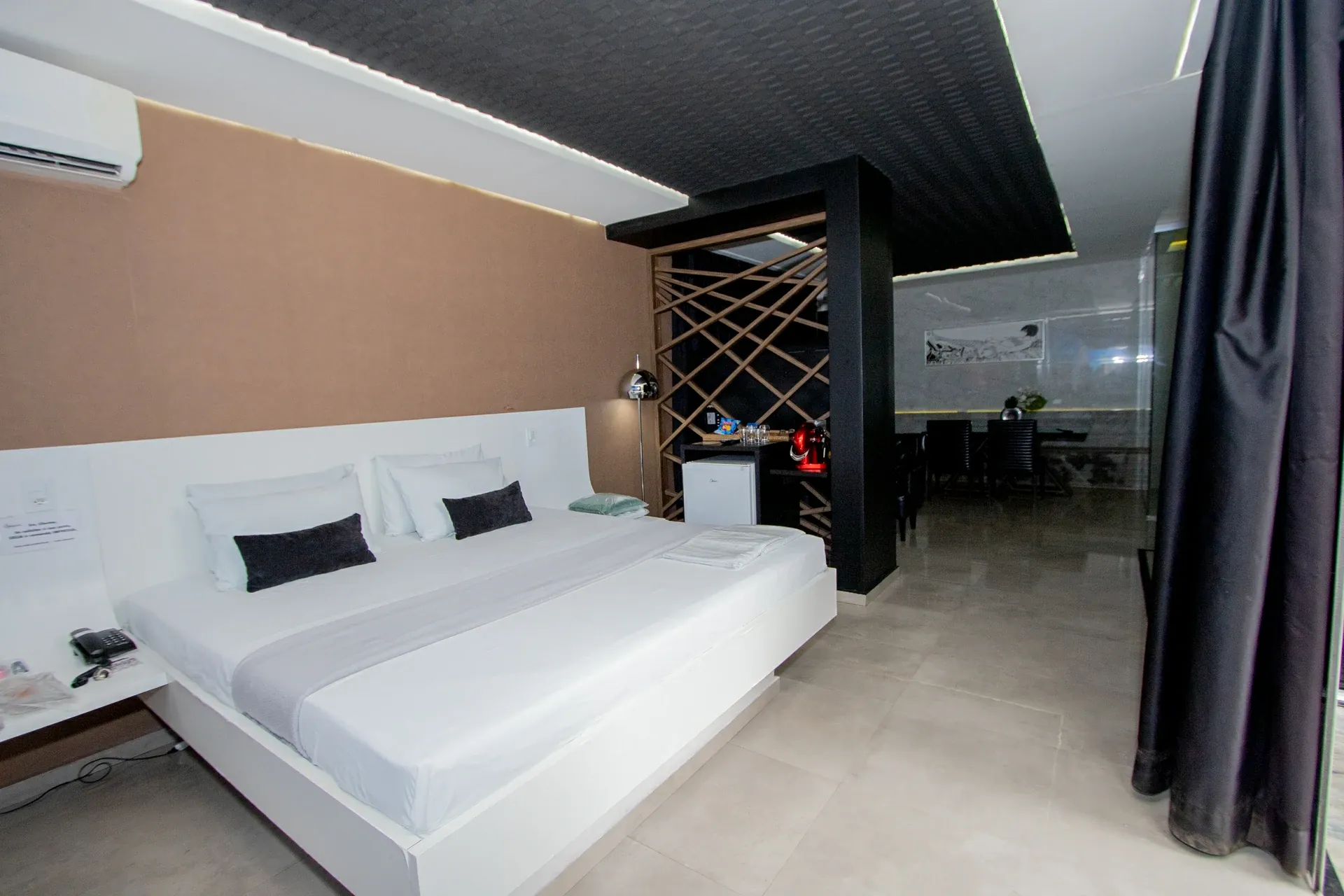 Suíte Opium do Motel Opium em Boa Vista: Vista lateral de suíte moderna com cama king branca de base flutuante, cabeceira branca contra parede acolchoa