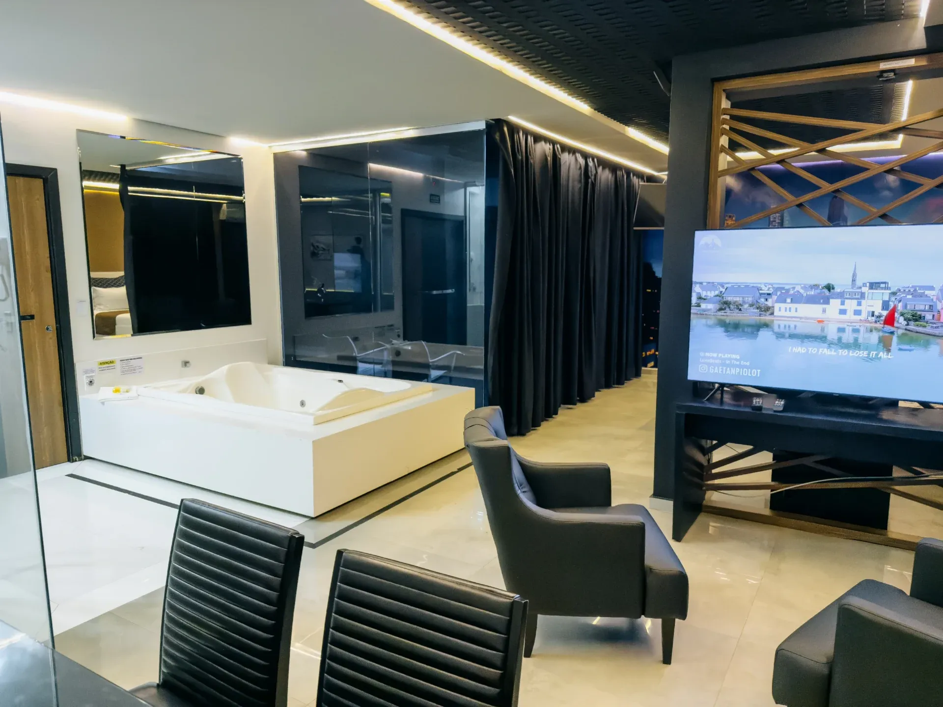 Suíte Opium do Motel Opium em Boa Vista: Visão ampla de suíte premium com banheira de hidromassagem dupla branca quadrada sobre plataforma central, TV 