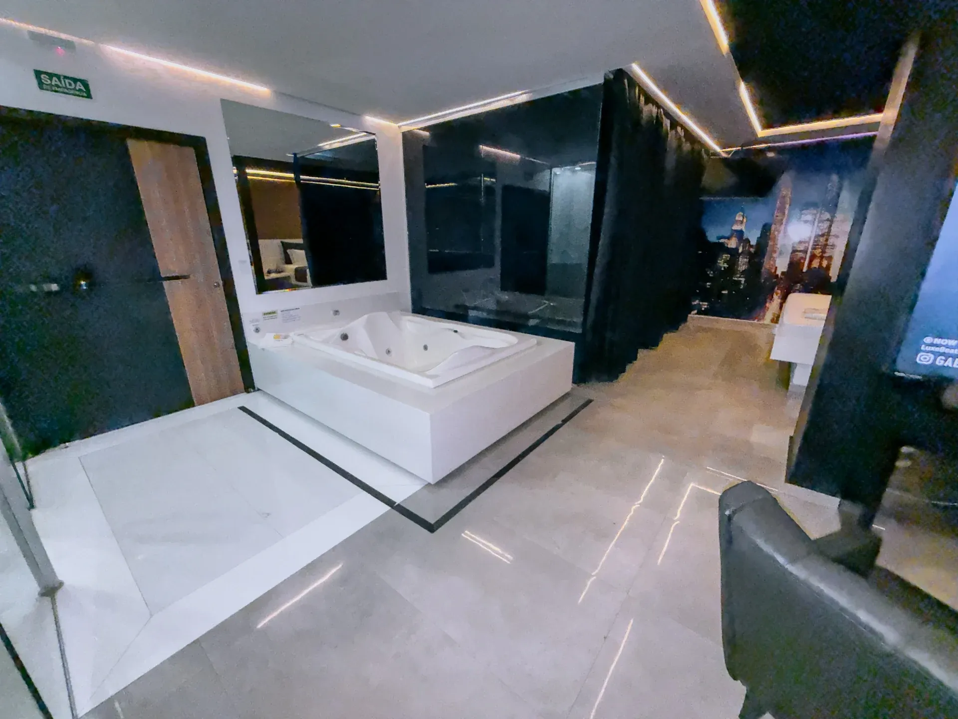 Suíte Opium do Motel Opium em Boa Vista: Mesma suíte com hidro dupla vista de outro ângulo, com mural de skyline ao fundo à direita e poltrona escura e