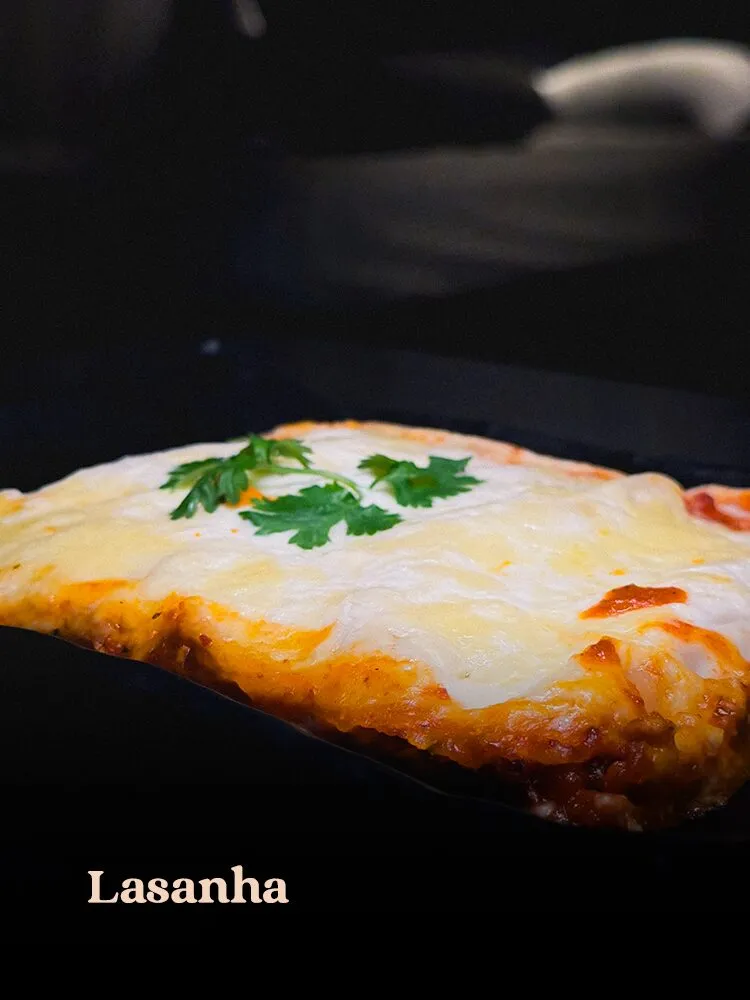 Fatia de lasanha com queijo gratinado e coentro sobre prato preto, em cena low-key com fundo quase totalmente · cardápio do Motel Opium