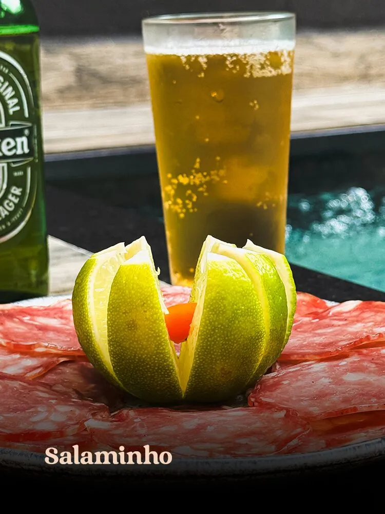 Prato de salaminho fatiado com limão cortado em flor ao centro, copo de cerveja e garrafa de Heineken, fotogra · cardápio do Motel Opium