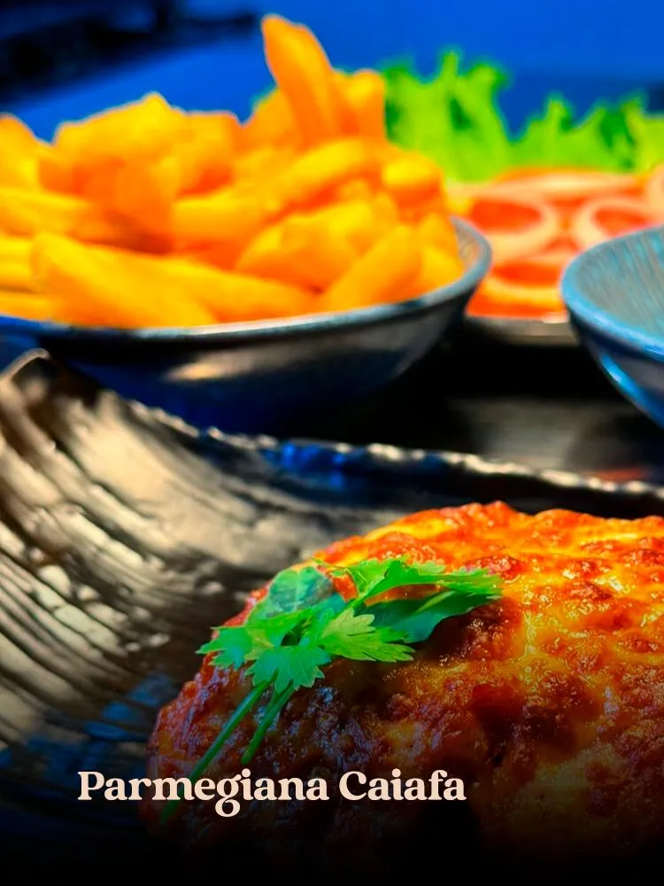 Close de frango à parmegiana gratinado com coentro em travessa preta texturizada, com fritas superssaturadas e · cardápio do Motel Opium