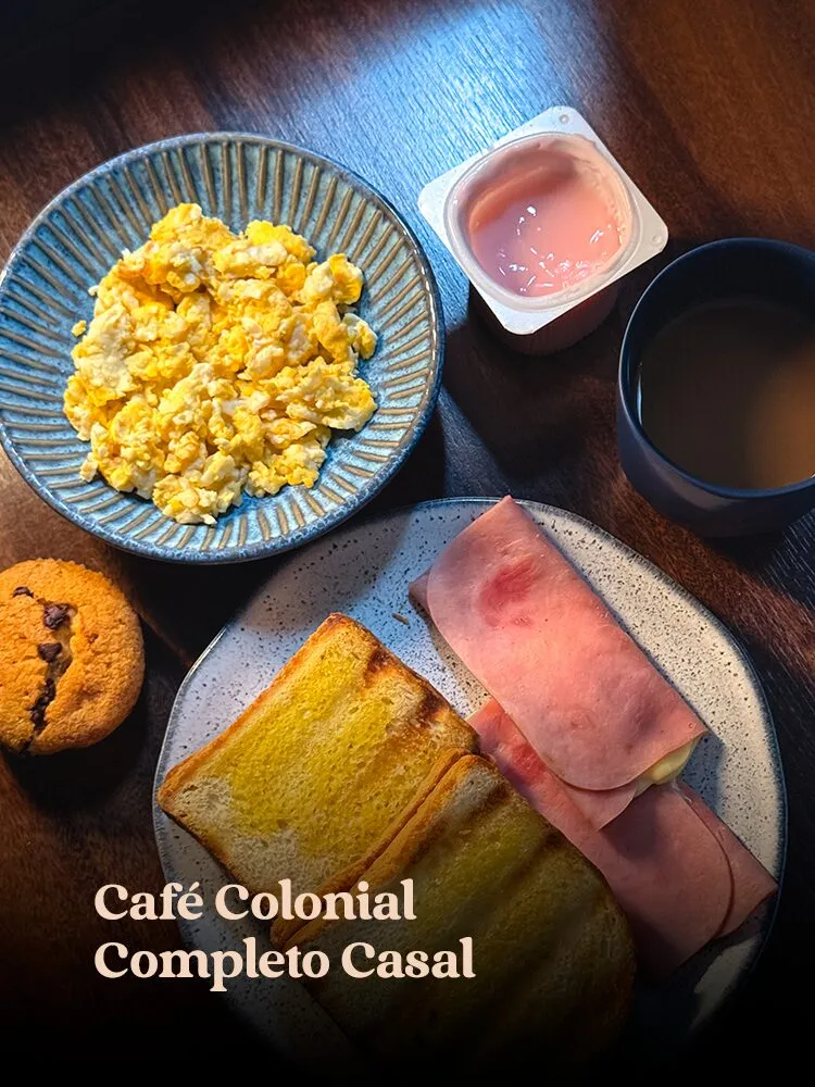 Café da manhã visto de cima em mesa de madeira escura: ovos mexidos em bowl azul canelado, torradas com presun · cardápio do Motel Opium