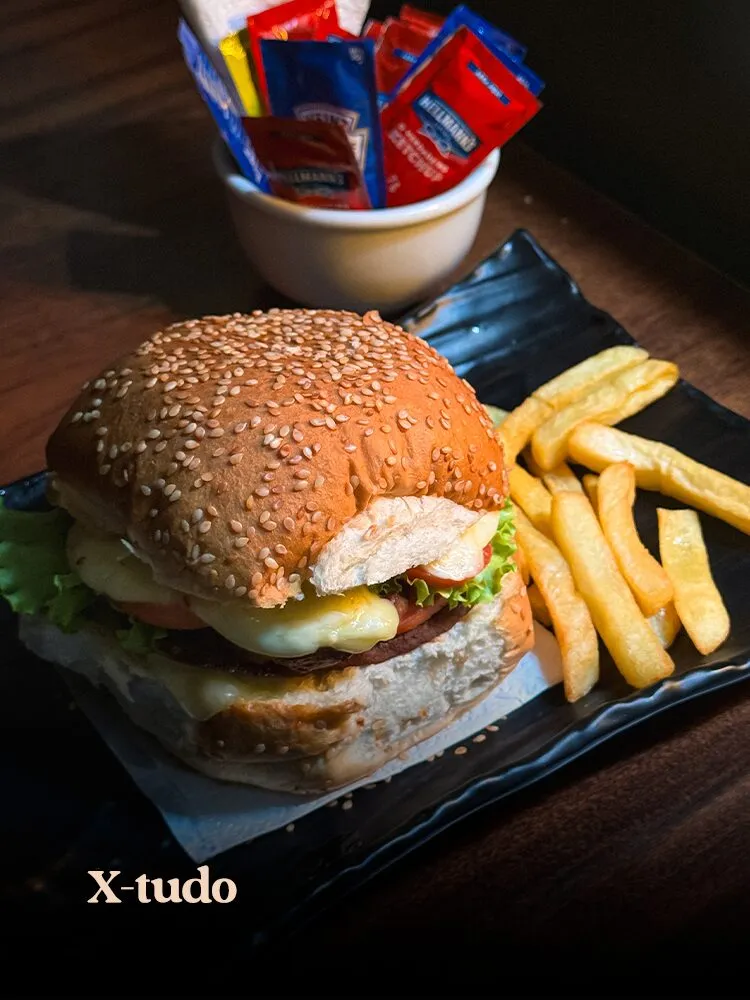 X-tudo com pão de gergelim e batatas fritas servidos em prato de ardósia preto sobre mesa de madeira · cardápio do Motel Opium