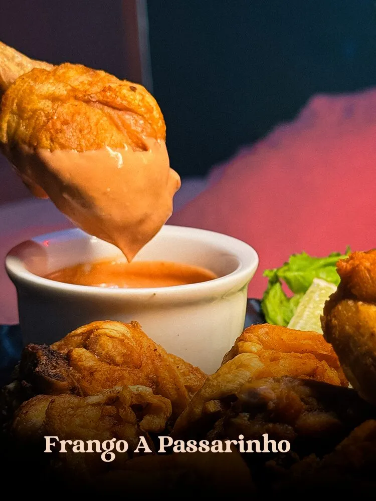 Pedaço de frango a passarinho sendo mergulhado em ramekin branco de molho rosé, com porção de frango frito e a · cardápio do Motel Opium