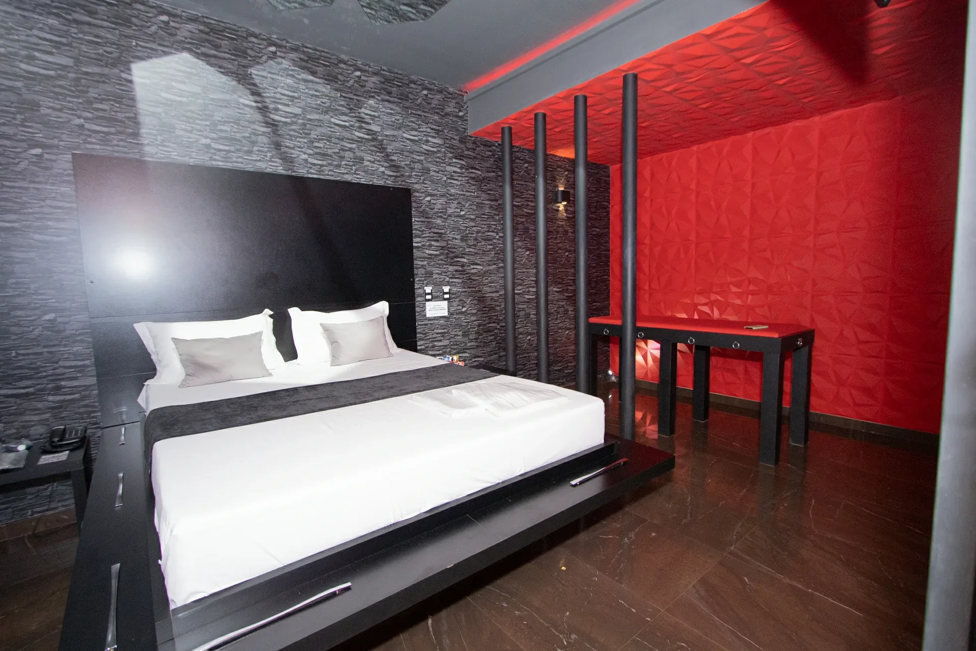 Suíte Sado do Motel Opium em Boa Vista: Plataforma preta flutuante com roupa branca e xale cinza, cabeceira preta alta contra parede de pedra cinza, q