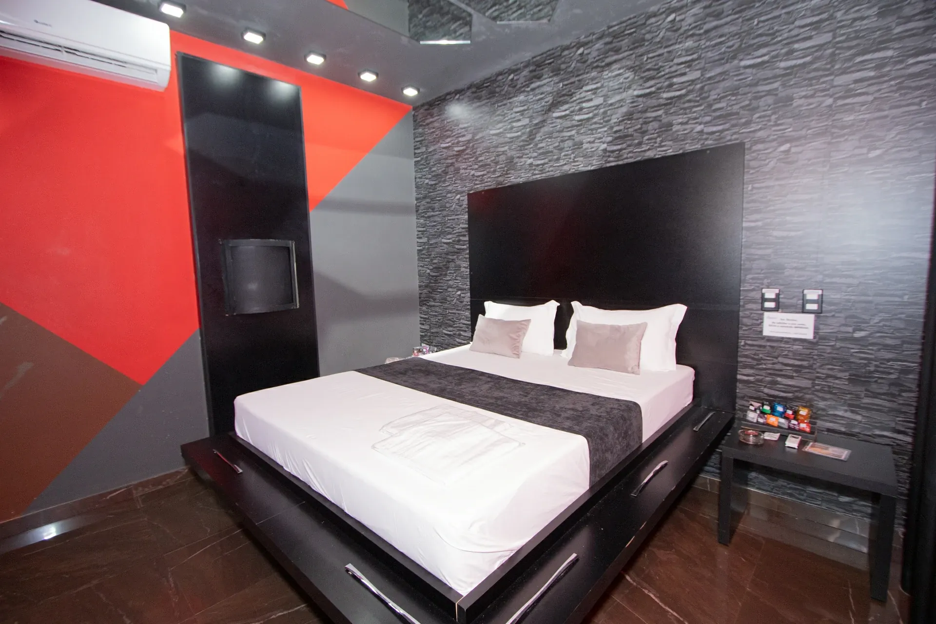 Suíte Sado do Motel Opium em Boa Vista: Cama king de plataforma preta centralizada, cabeceira em painel preto sobre parede de pedra cinza, parede late