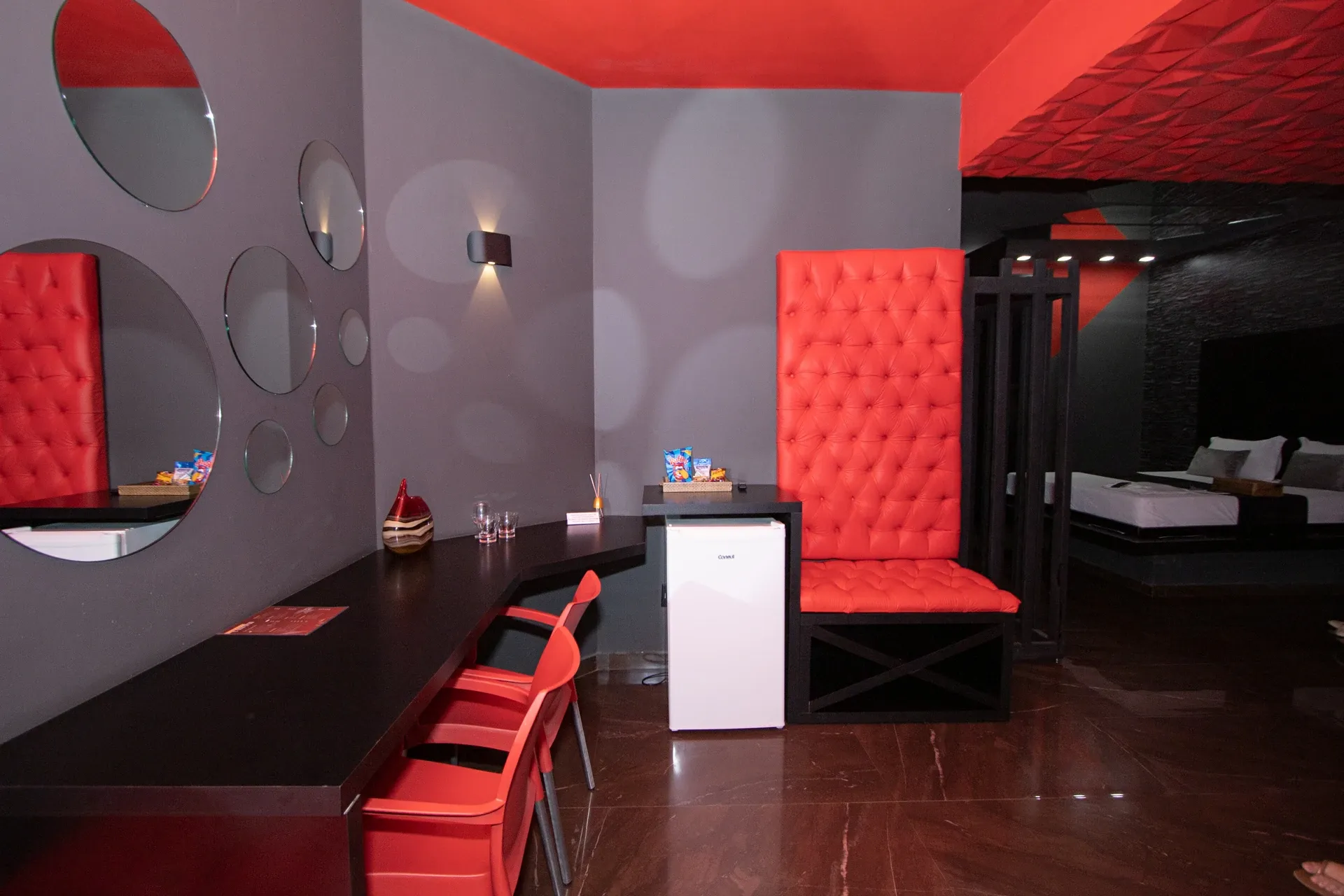 Suíte Sado do Motel Opium em Boa Vista: Área de estar/copa da suíte Sado: bancada preta em L sob composição de espelhos redondos na parede cinza, duas