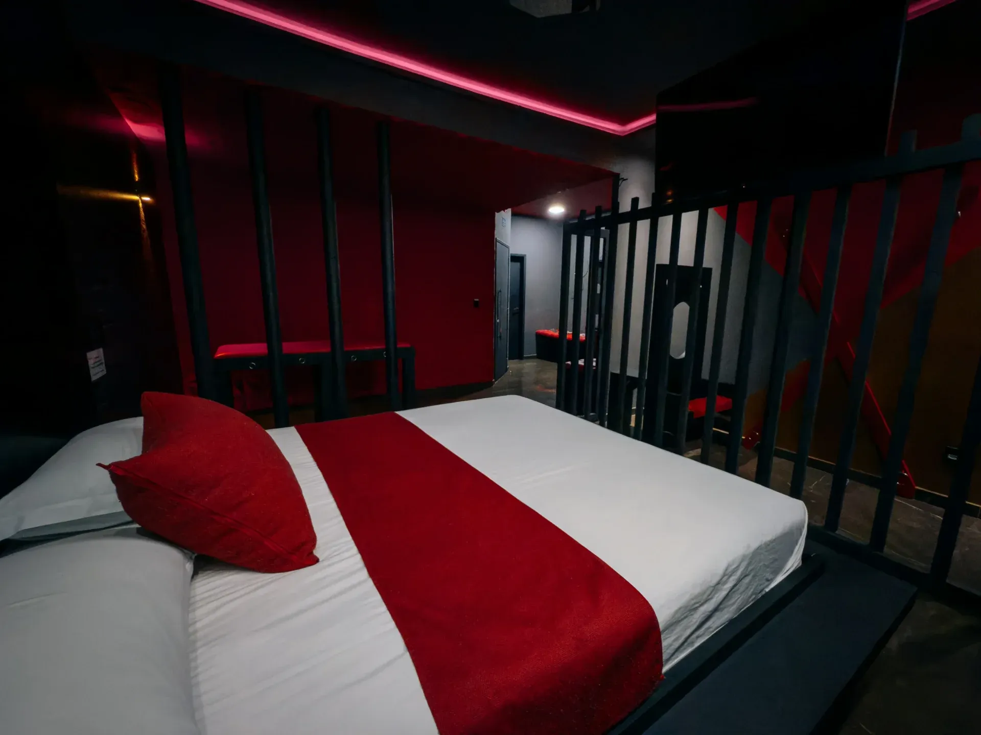 Suíte Sado do Motel Opium em Boa Vista: Suíte temática dark/BDSM vista por trás de grades pretas estilo cela: cama com faixa vermelha e almofada verme