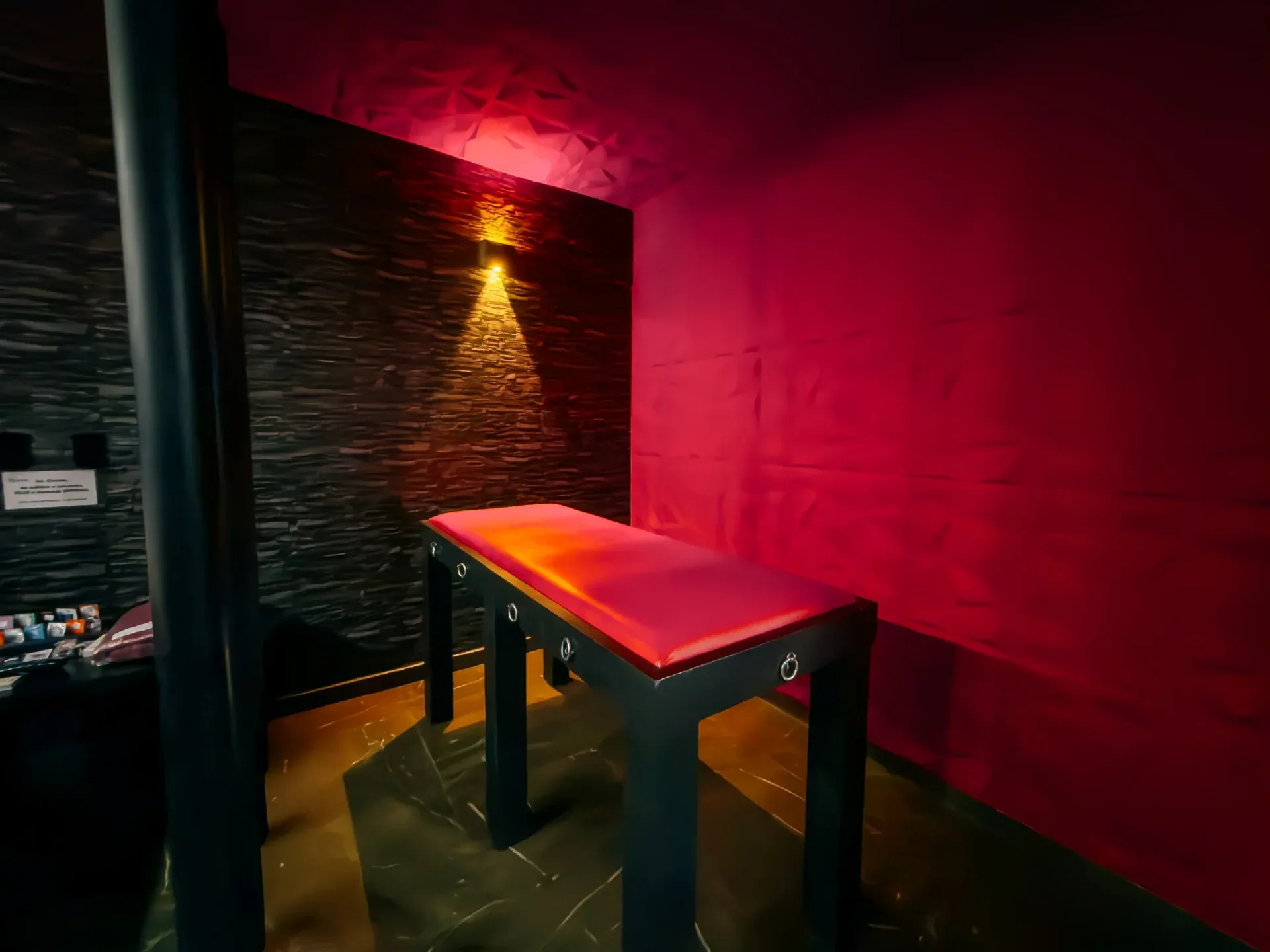 Suíte Sado do Motel Opium em Boa Vista: Mesma suíte dark em diagonal: cama de casal com faixa e almofada vermelhas em primeiro plano, parede de pedra 
