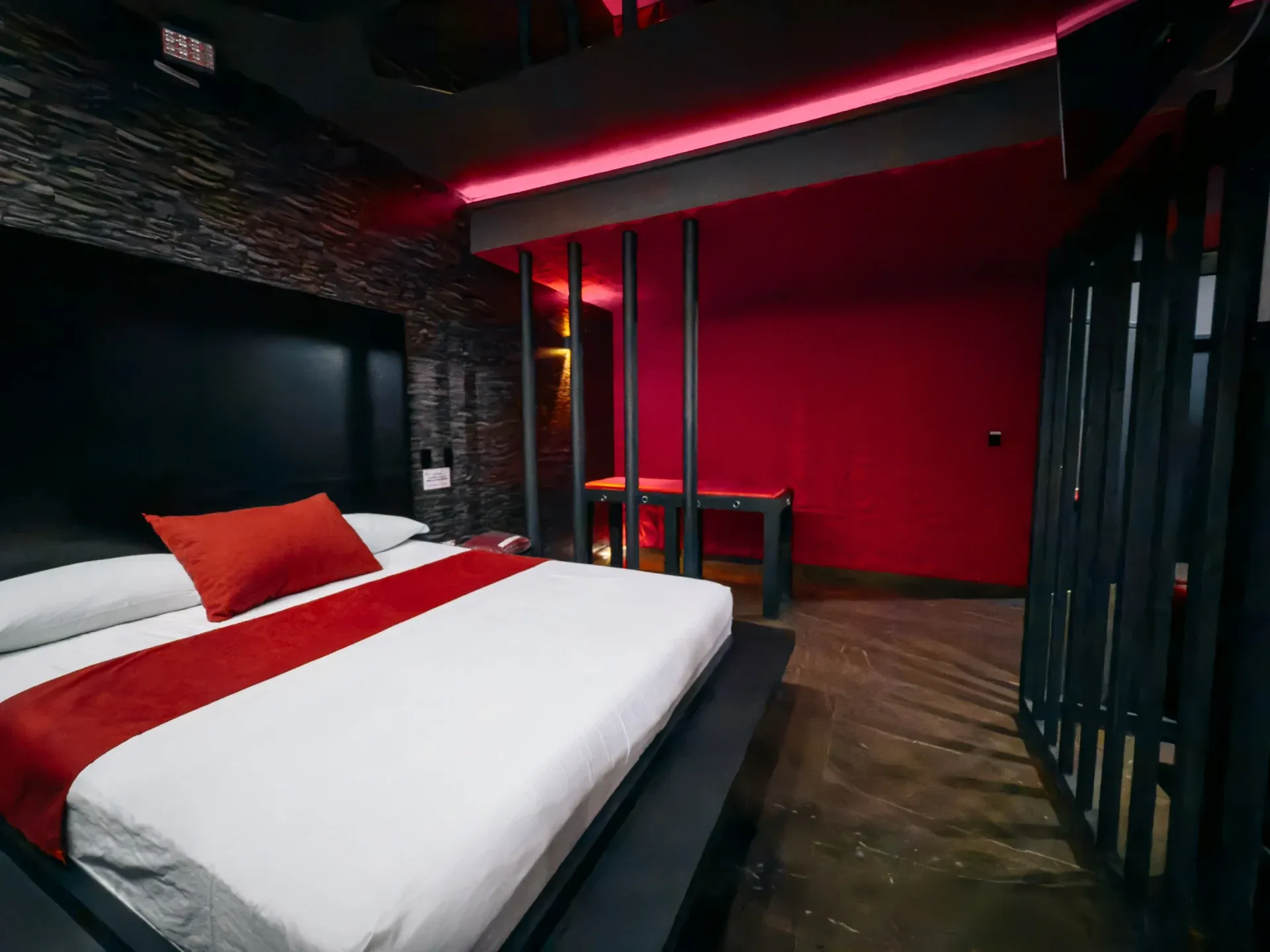 Suíte Sado do Motel Opium em Boa Vista: Parede geométrica em vermelho, preto, cinza e ocre atrás da cama flutuante com peseira vermelha, banco erótico