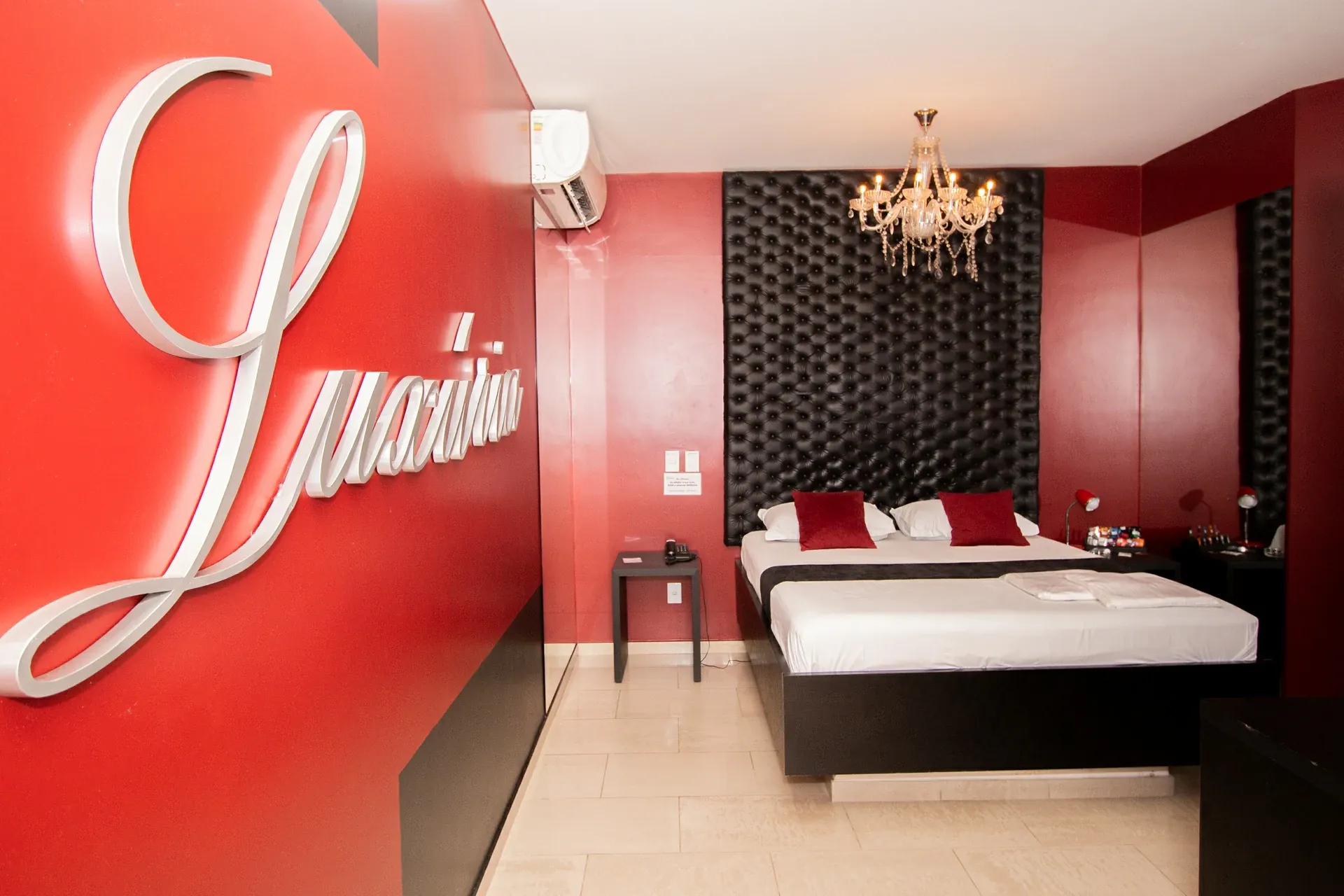Suíte VIP do Motel Opium em Boa Vista: Suíte Luxúria em vista diagonal: letreiro 3D prateado em caligrafia cursiva 'Luxúria' dominando a parede verme