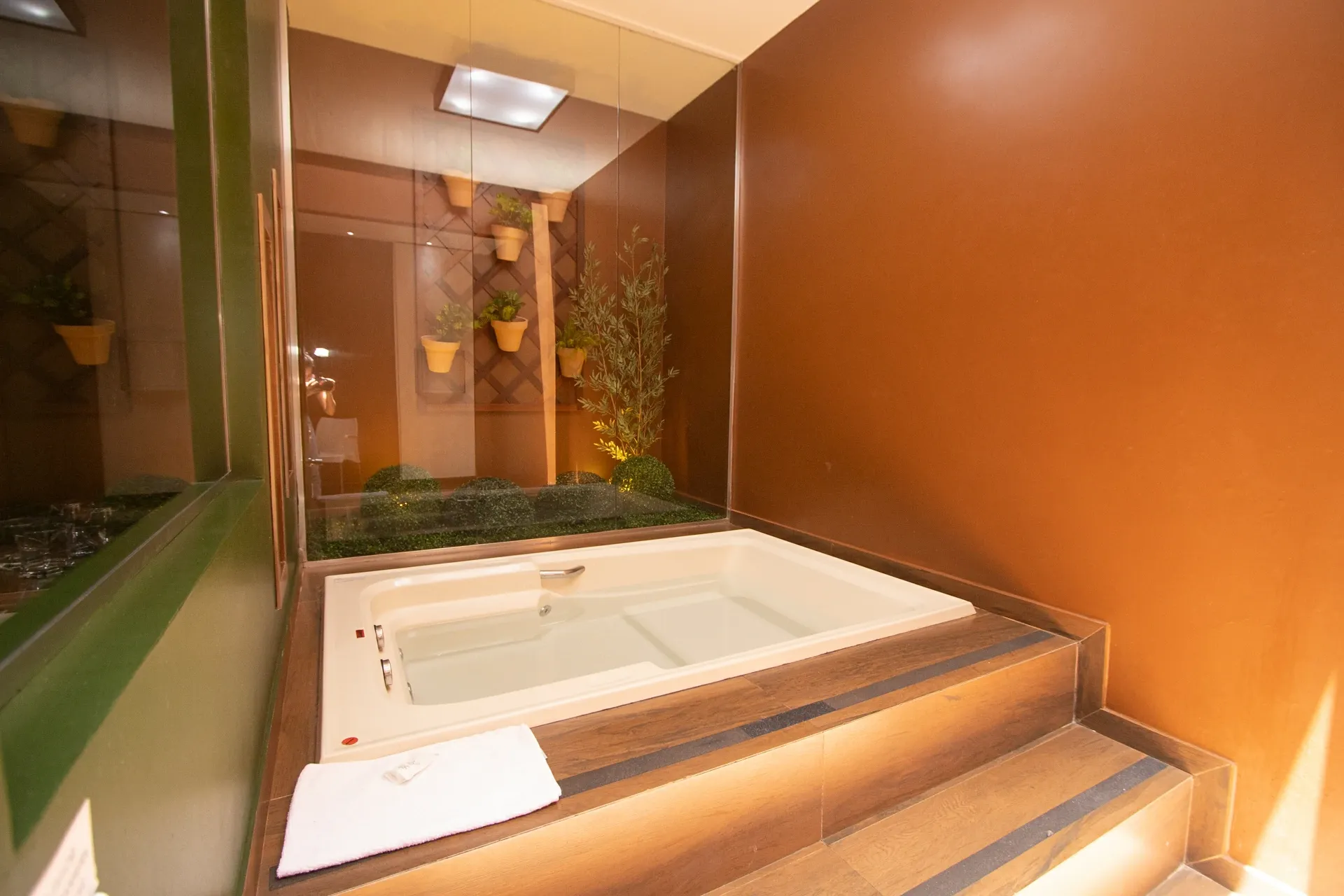 Suíte VIP do Motel Opium em Boa Vista: Banheira de hidromassagem branca embutida em plataforma com degraus revestidos de madeira e iluminação embutid
