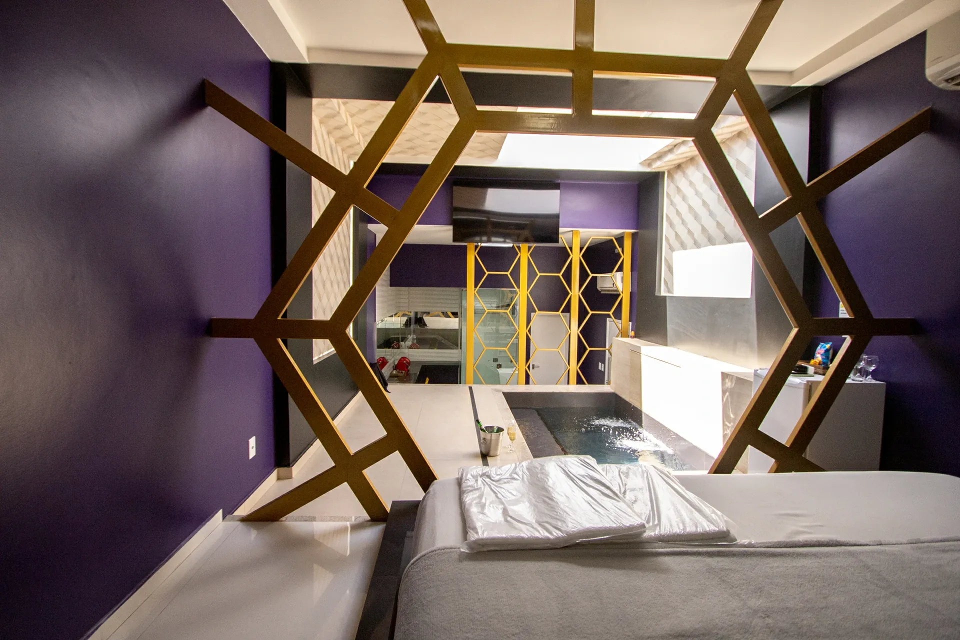 Suíte VIP Plus do Motel Opium em Boa Vista: Vista da cama em direção à piscina de hidromassagem, enquadrada através da treliça dourada hexagonal que emold